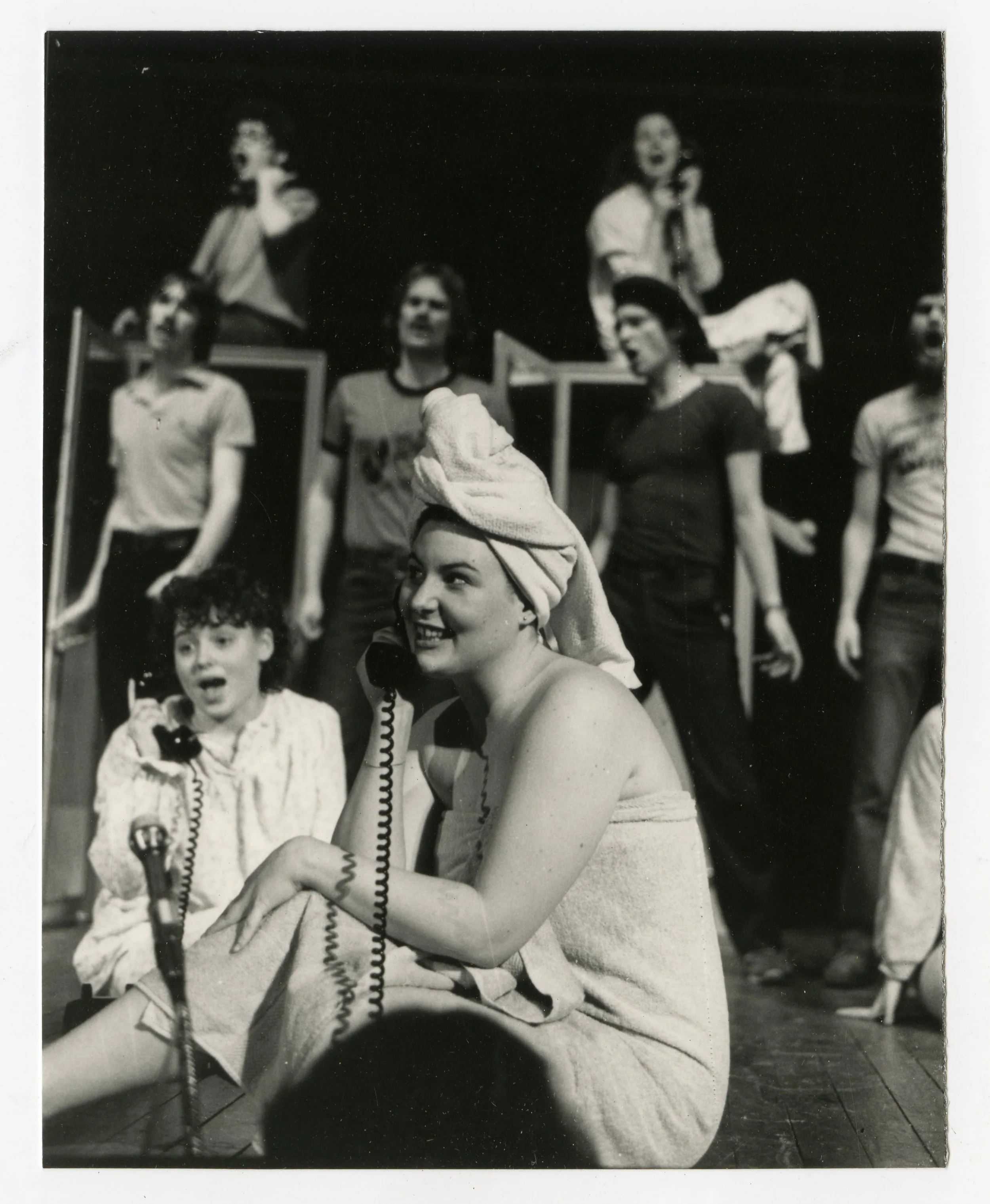 Black-and-white photo of women in a theatrical rehearsal, some sitting, some standing, with two women sitting on the floor talking on telephones, one woman with a towel wrapped around her head.
