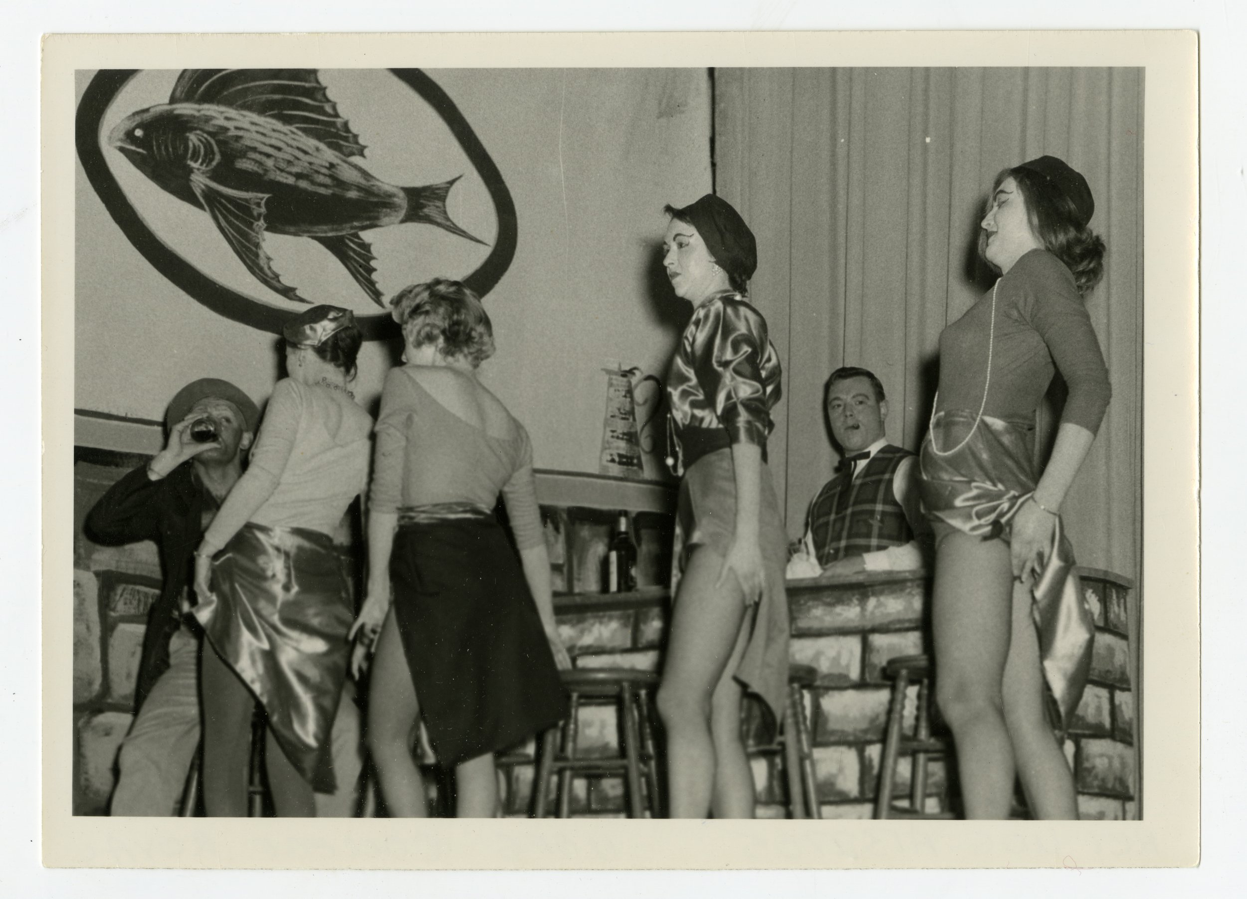 Black and white photo of a bartenders' show with women dressed in shiny, revealing outfits standing on bar stools.