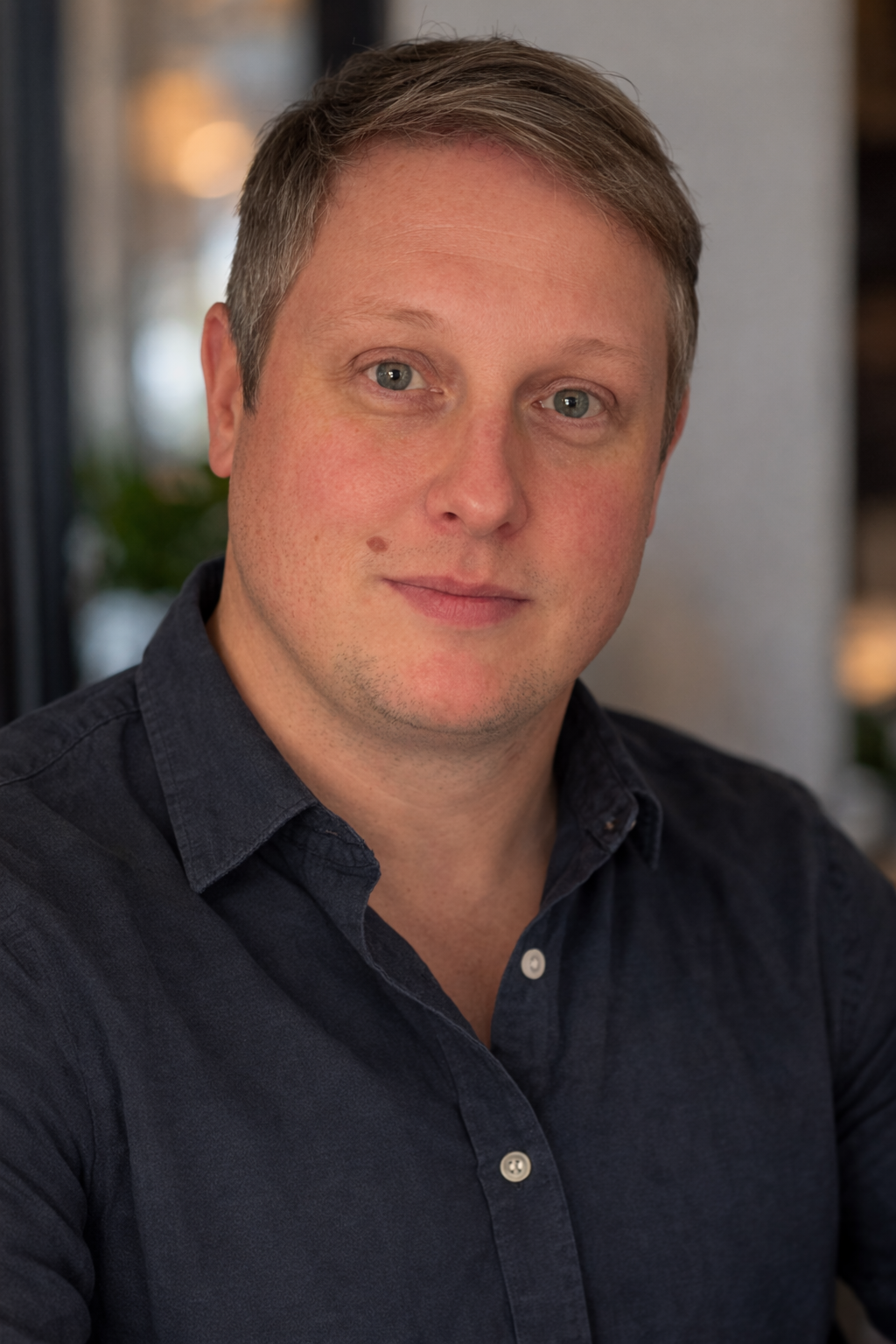 Close-up portrait of a man with short light brown hair, fair skin, blue eyes, wearing a dark button-up shirt, in an indoor setting with blurred background.