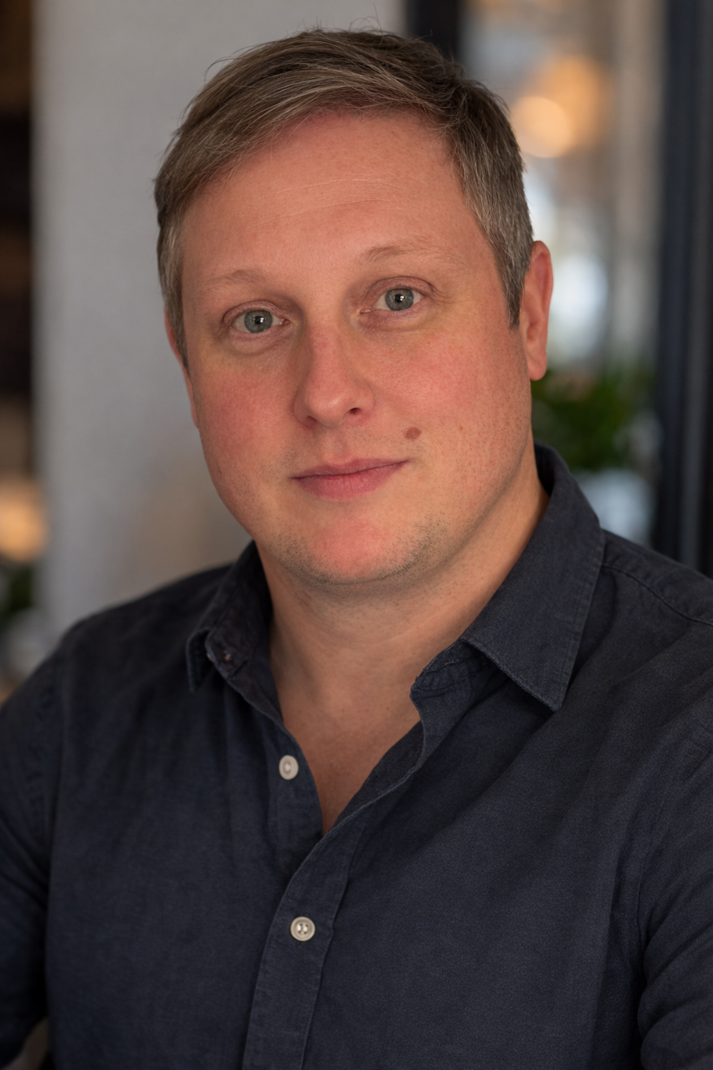 A portrait of a man with fair skin, short light brown hair, blue eyes, and wearing a black collared shirt.