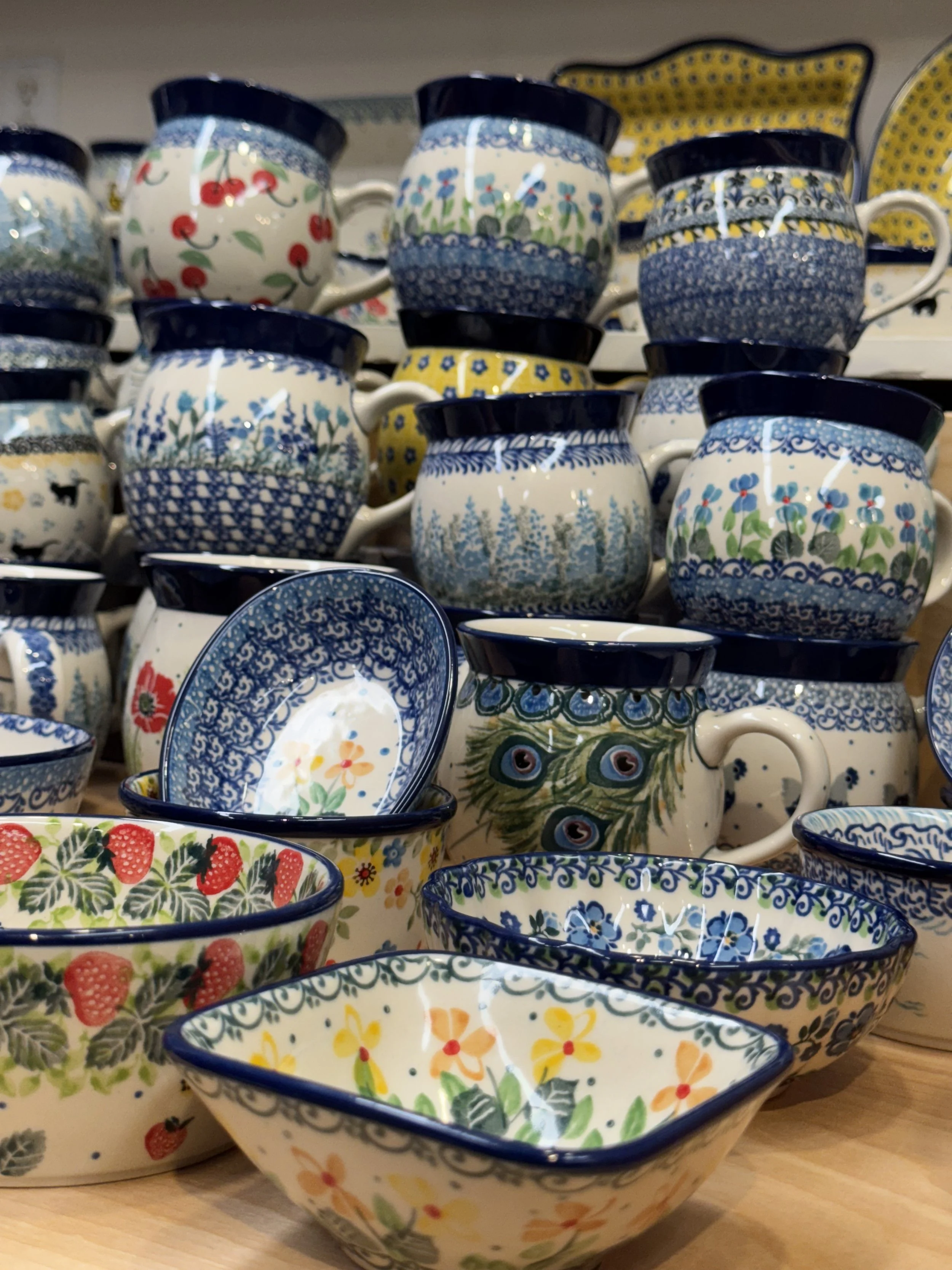 Stacked ceramic mugs, Polish Pottery, bowls, and dishes with floral, berry, and peacock feather patterns in blue, white, red, yellow, and green colors, displayed on a wooden surface.