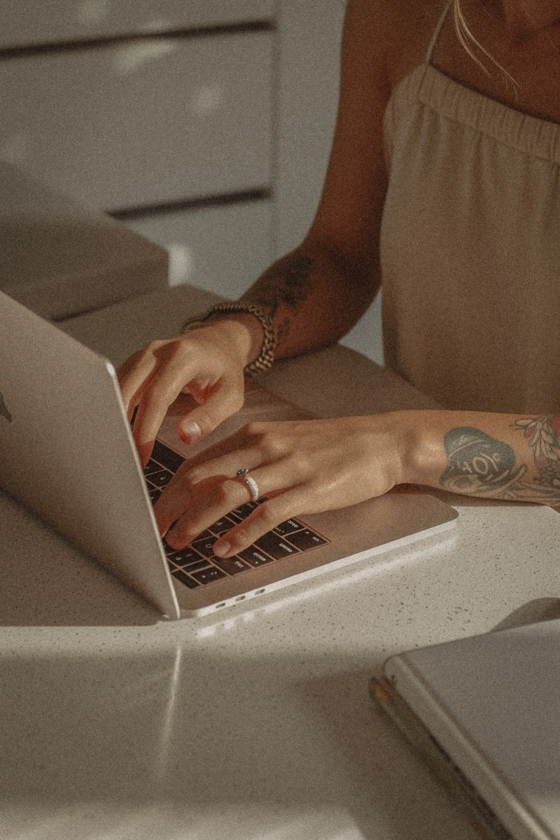 Person working on a laptop at a table, with tattoos on their arms and wearing a ring and a bracelet.