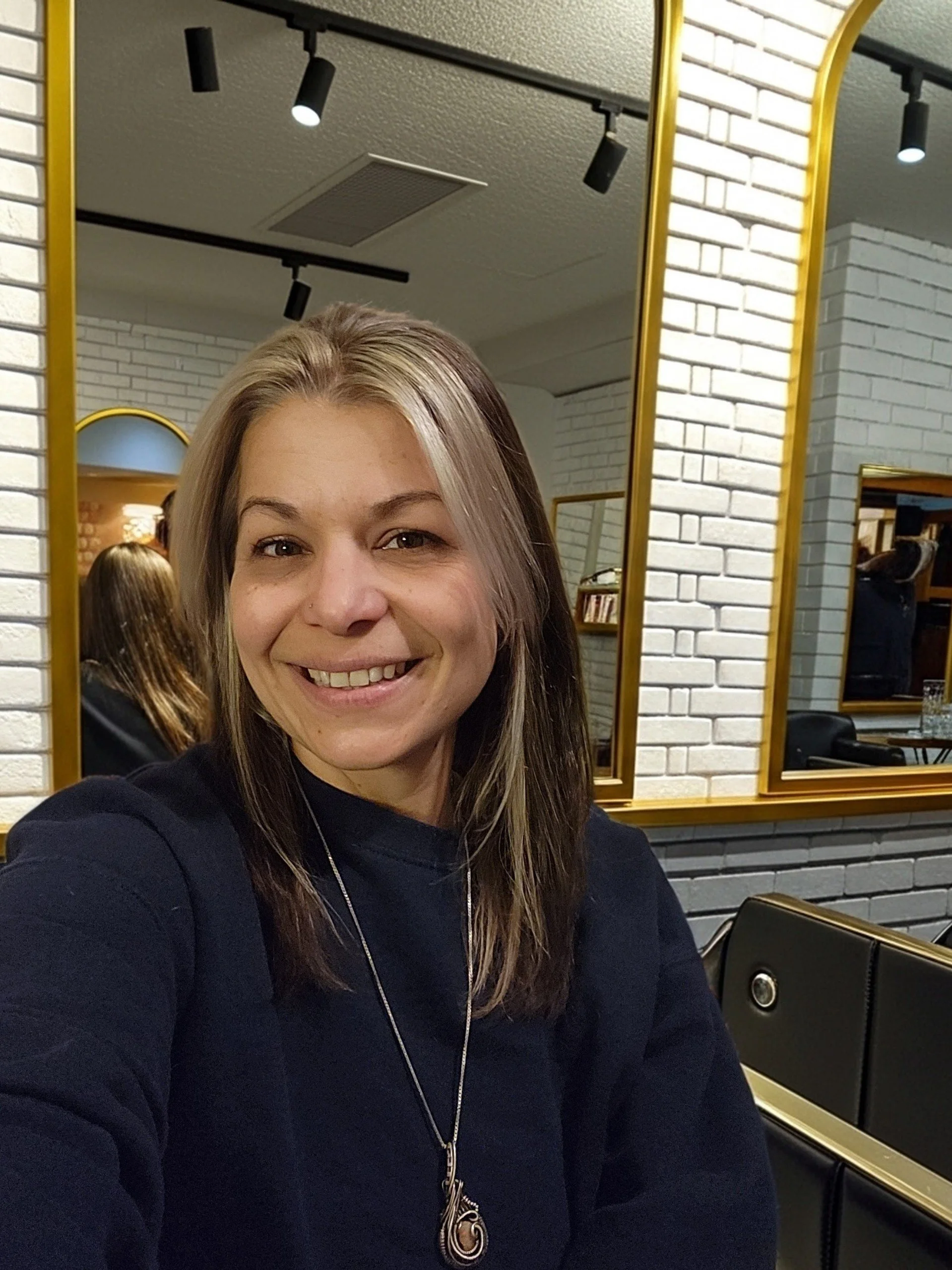 A woman with shoulder-length brown hair and blonde highlights smiling at the camera, sitting in a room with white brick walls, gold-framed mirrors, and black and gold furniture.