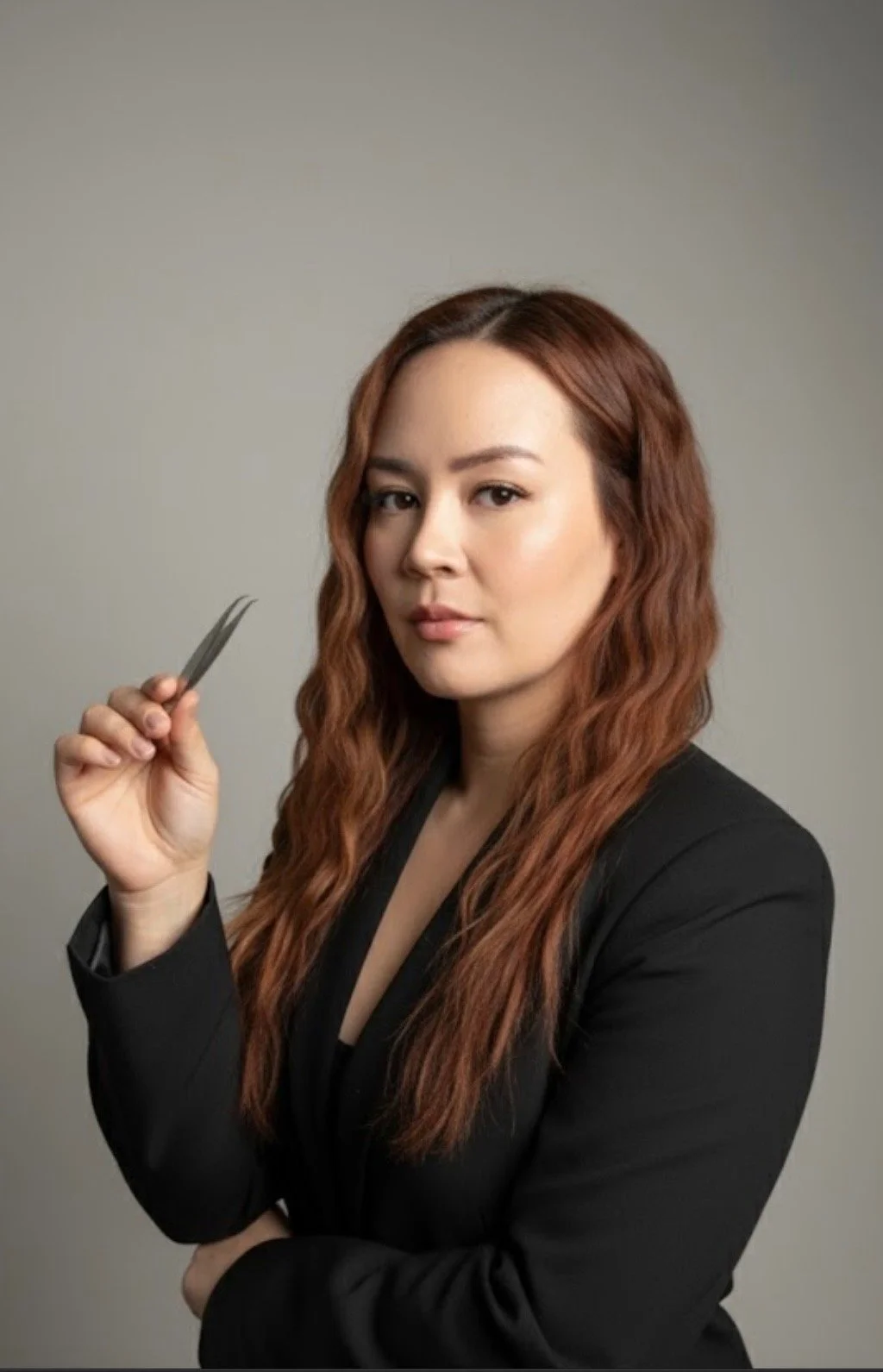 A woman with long red hair holding a pair of tweezers, wearing a black blazer, against a plain neutral background.