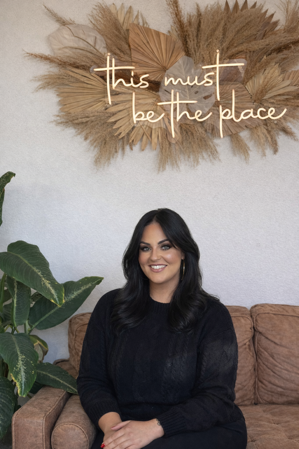 Smiling woman with dark hair and hoop earrings sitting on a brown couch, with a green plant beside her and a wall decoration that reads 'this must be the place' in neon script above her.