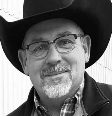 A man wearing glasses, a cowboy hat, and a checkered shirt indoors.