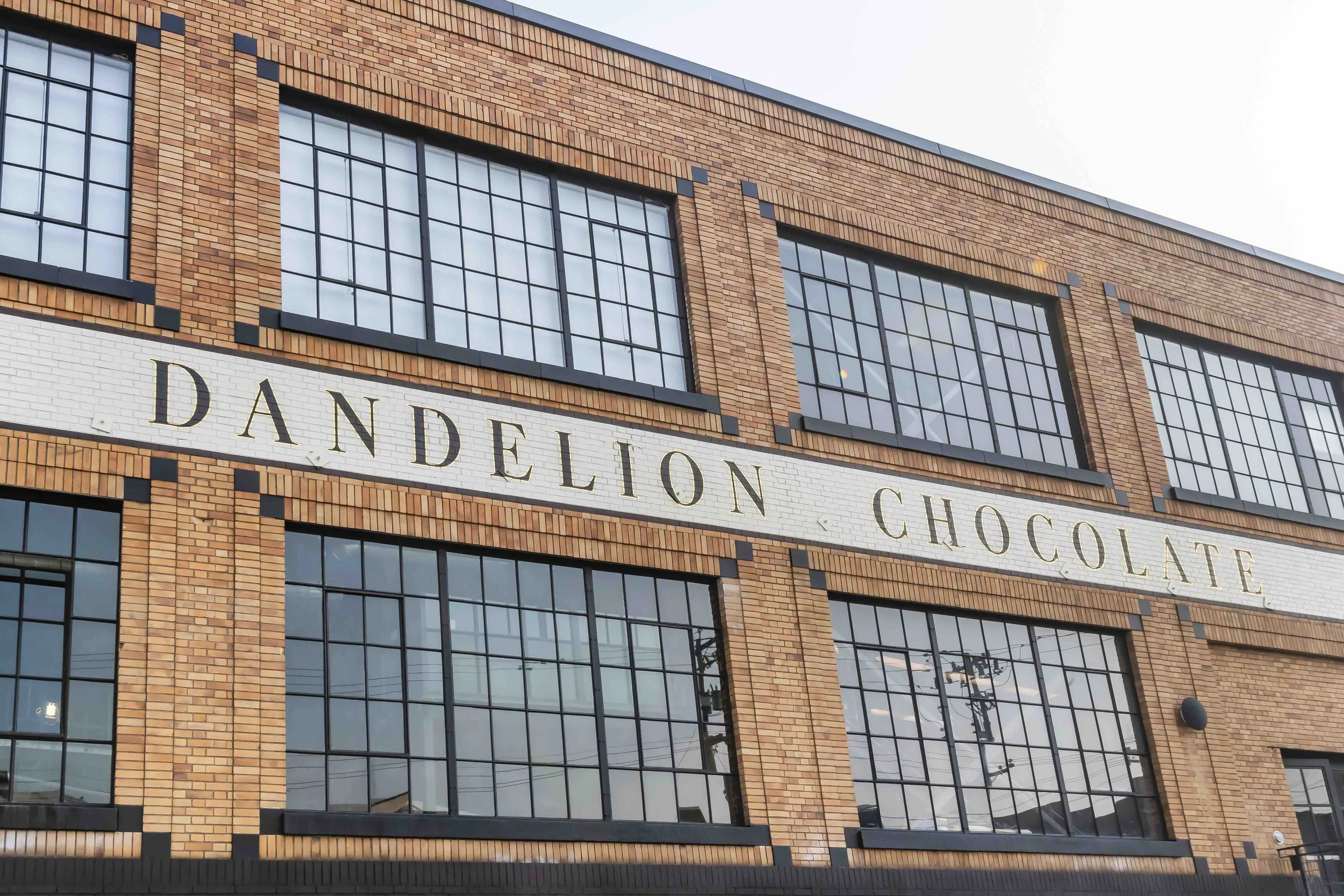 Facade of a brick building with large black-frame windows and a white sign reading 'Dandelion Chocolate'