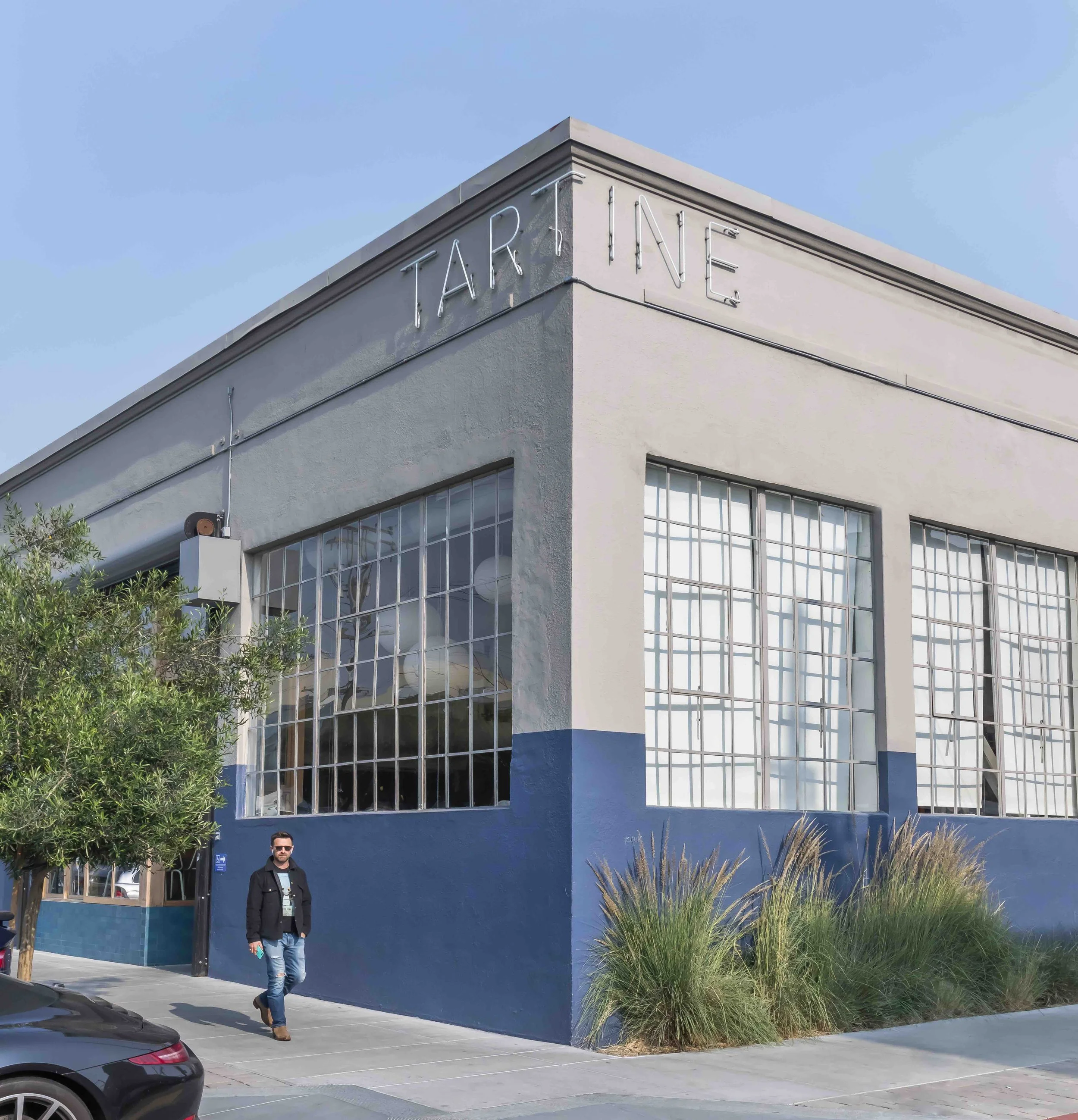 Corner view of a modern commercial building with large grid windows, blue and off-white facade, and neon text on top reading 'TARTINE' and 'INE'. A man in sunglasses, black jacket, and jeans walks on sidewalk in front of the building, with greenery a