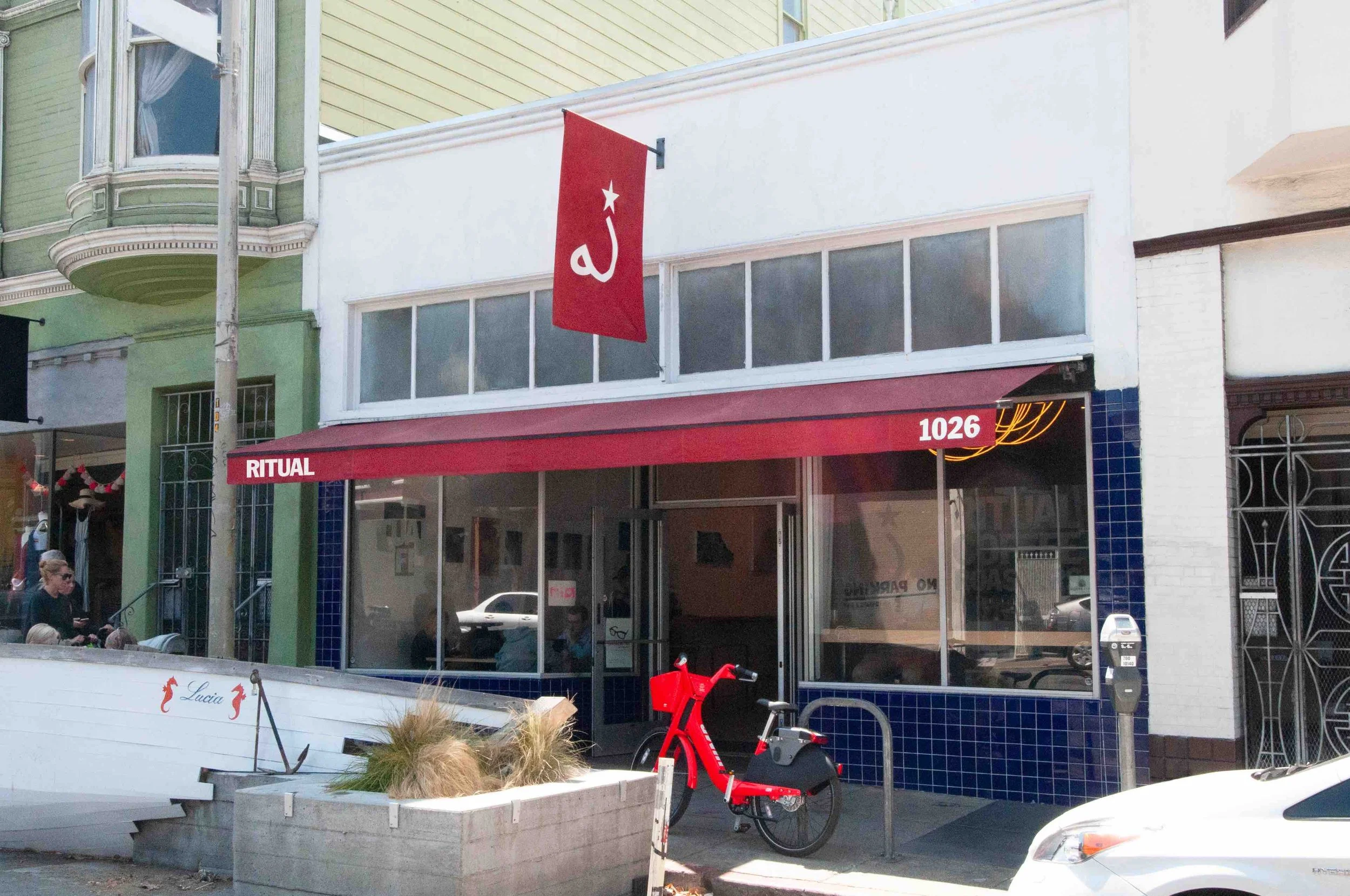 Facade of a building with a red awning labeled 'RITUAL' and the number 1026, blue tiled trim, a red flag with a white star and symbol, large glass windows, a red bike parked outside, and people sitting and walking nearby.