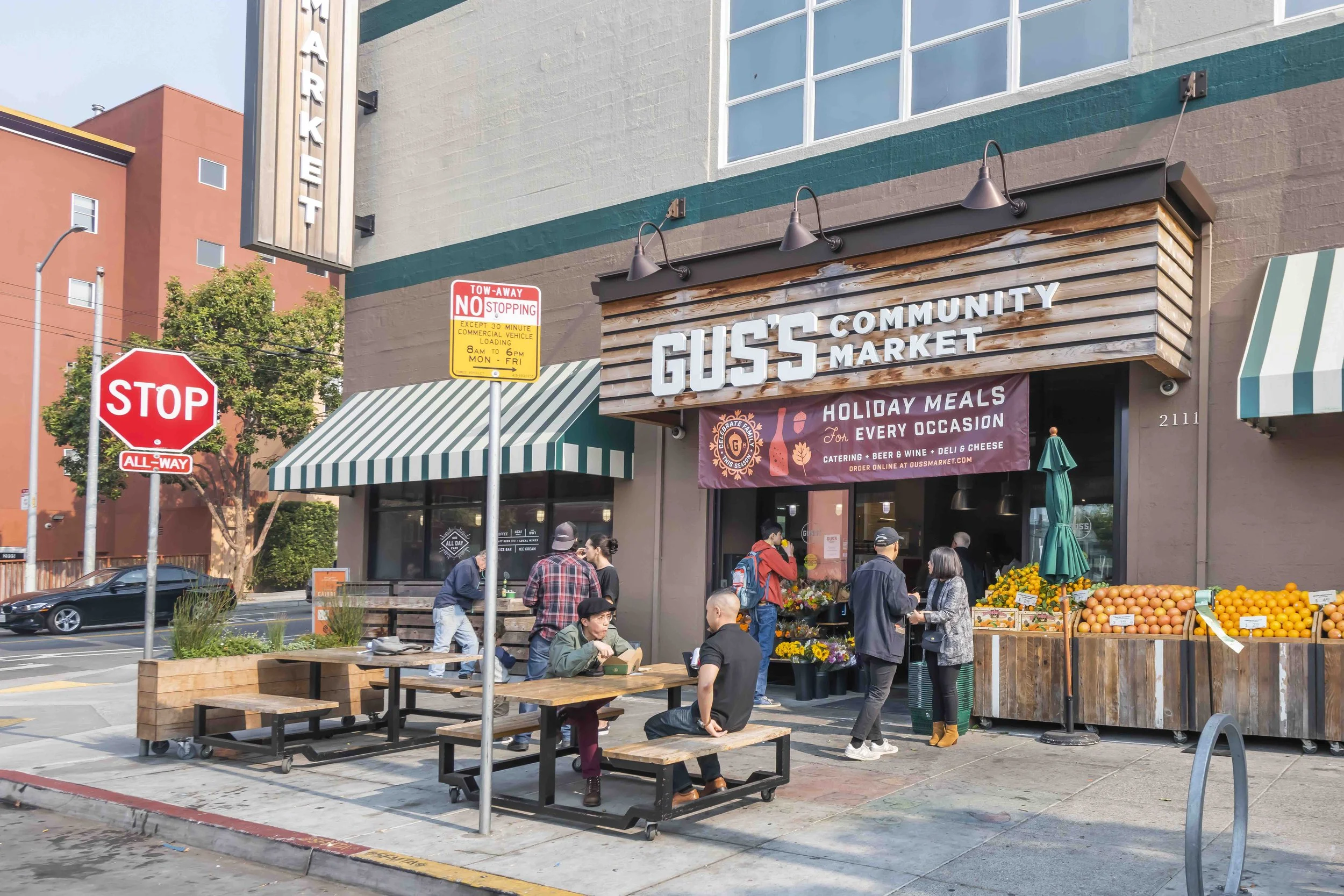 People outside Guss Community Market, a grocery store with a sign that reads 'Holiday Meals for Every Occasion,' offering catering, beer, wine, deli, and cheese. There are outdoor tables with people sitting, a fruit stand with oranges, and pedestrian