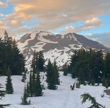 Photo of South Sister Mountain around Bend, Oregon. A snow-covered volcano with patches of snow on rocky slopes and a forest of evergreen trees in the foreground under a partly cloudy sky. Photo taken by Luc Gensler.