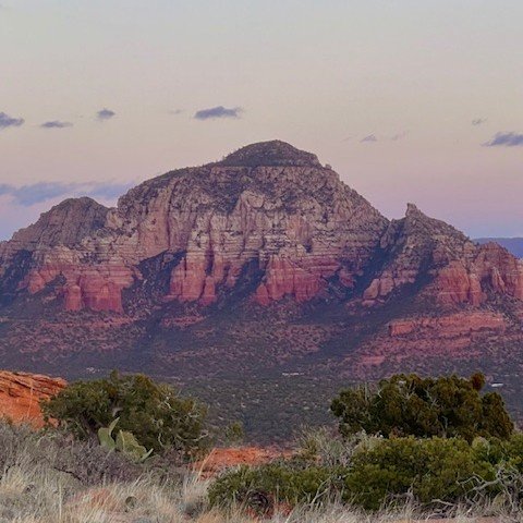 Photo of Capital Butte in Sedona, Arizona. A large mountain with reddish and grayish rock formations, set against a cloudy sky, with desert vegetation in the foreground. Photo taken by Luc Gensler.