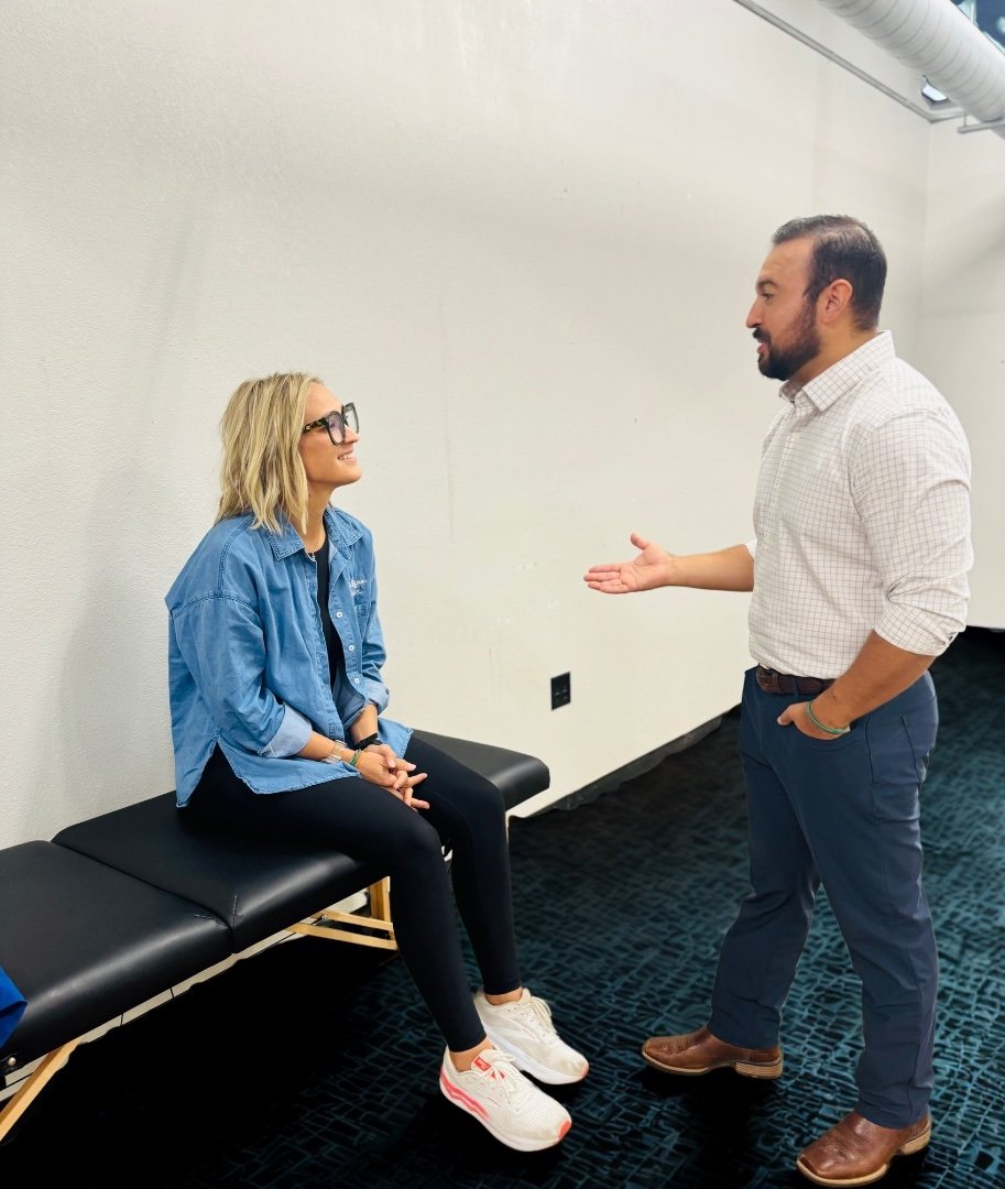 A woman with blonde hair, glasses, and casual clothes sitting on a black bench, talking to a man with dark hair, beard, and business casual clothes standing in front of her, inside a room with plain white walls and dark carpet.