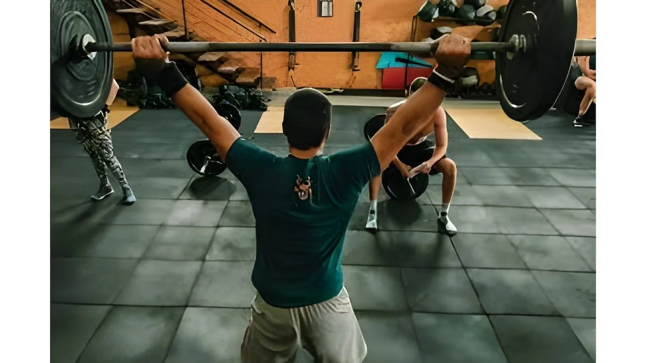A person lifting a barbell with weights overhead in a gym while others are sitting on the floor and preparing equipment in the background.