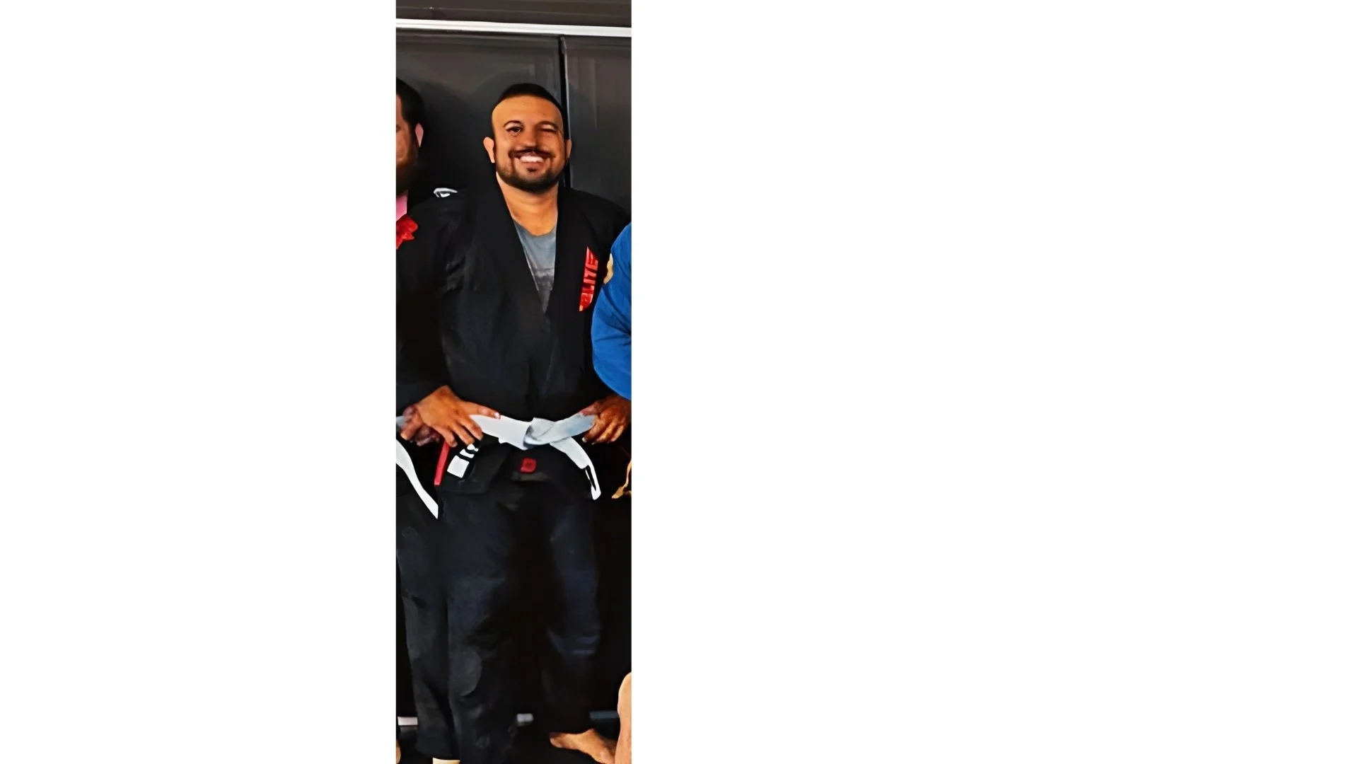Man wearing a black Brazilian jiu-jitsu gi with a white belt, smiling and standing indoors.