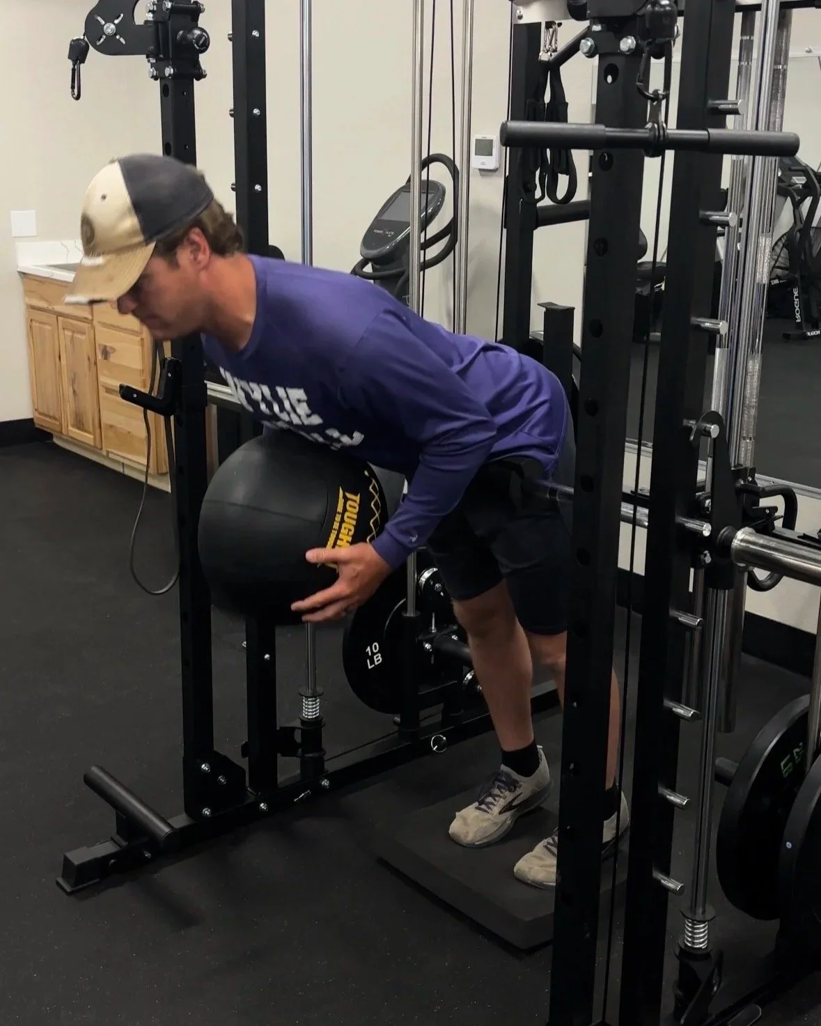A man in a baseball cap and athletic clothes performs a bent-over exercise with a medicine ball in a gym.