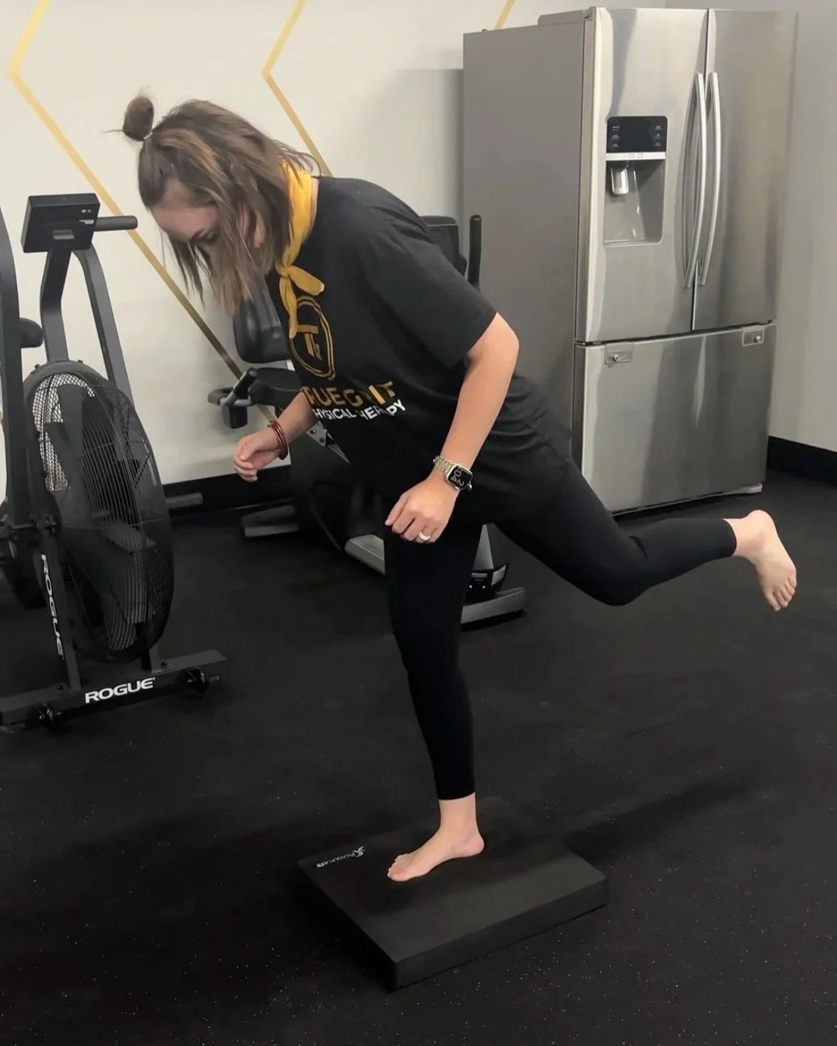 A woman performing a balance exercise on a black balance board in a gym, wearing black athletic clothing, a yellow scarf, and a smartwatch, with a refrigerator and gym equipment in the background.