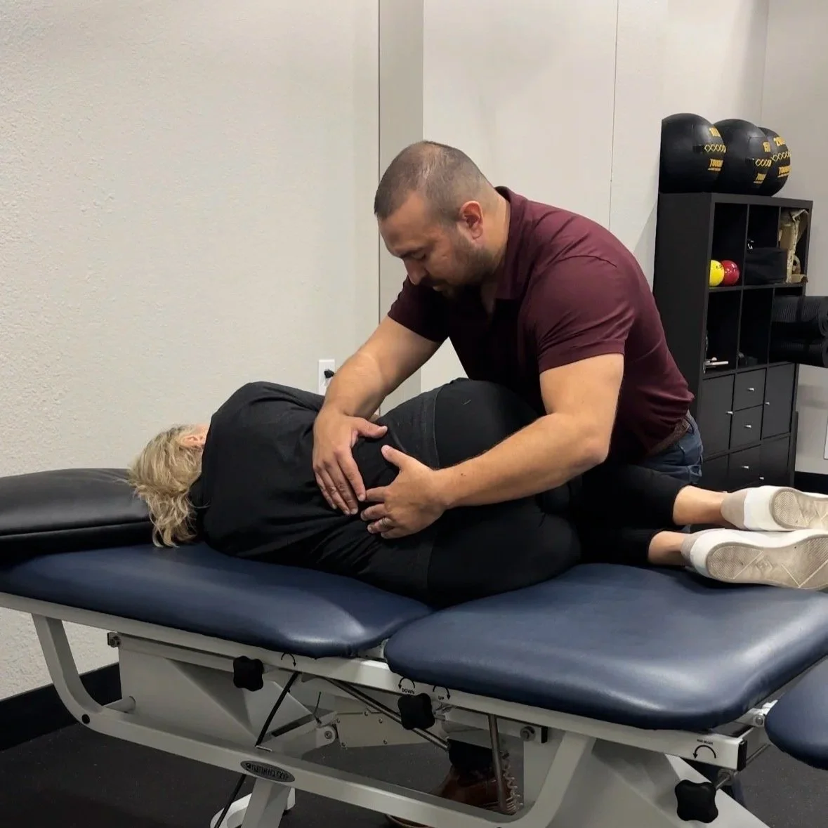 A man providing physical therapy or chiropractic care to a woman lying on a treatment table, holding her knee and hip area.