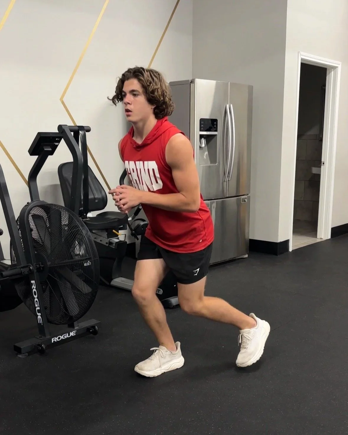 A young man in a red sleeveless shirt and black shorts running on a treadmill in a gym.