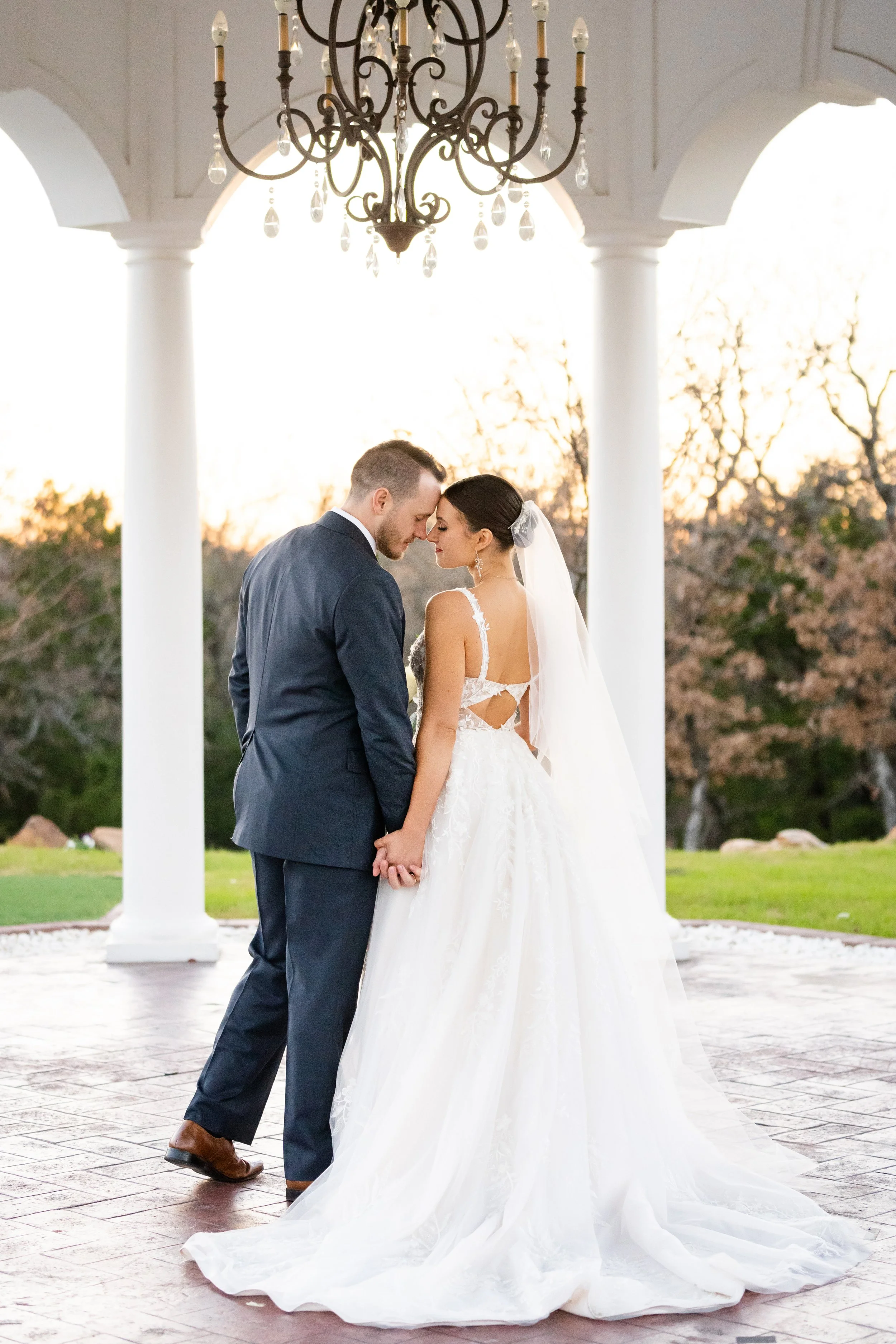 A bride and groom holding hands and touching foreheads in an outdoor wedding setting during sunset, under a white pavilion with a chandelier hanging above.