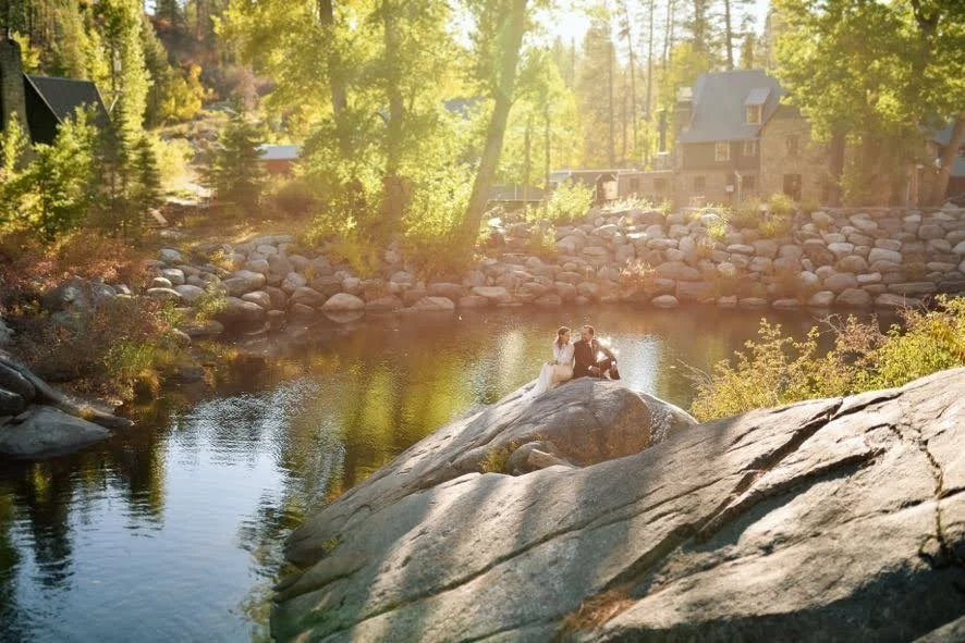 A couple dressed in wedding attire sitting on a large rock near a river in a wooded area, with rocky banks and a house visible in the background during sunset or golden hour.