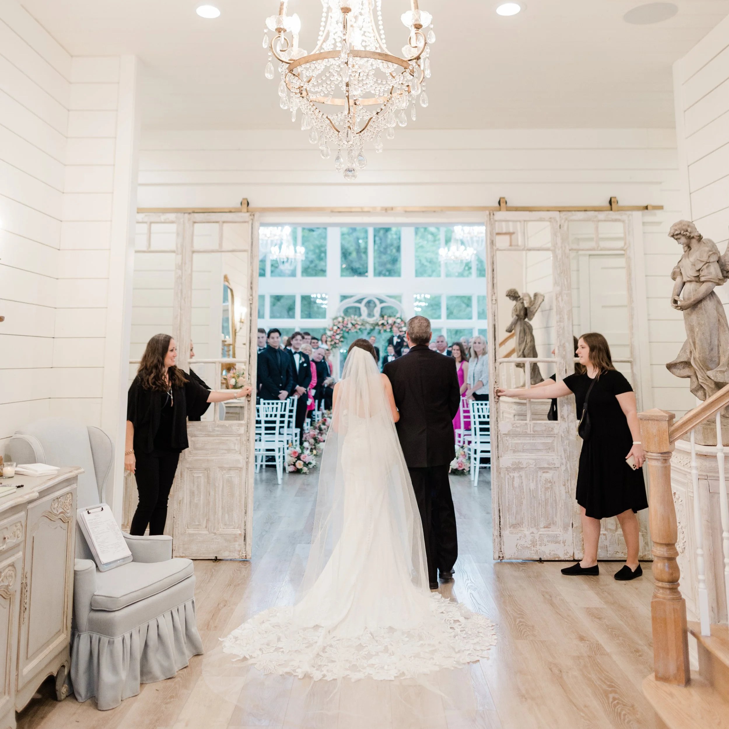 A bride and groom walk through open rustic wooden doors into a decorated wedding venue with guests awaiting inside. The bride wears a white wedding gown with a veil, and the groom wears a black suit. The venue has white walls, a chandelier, and statues, with floral decorations visible in the background.