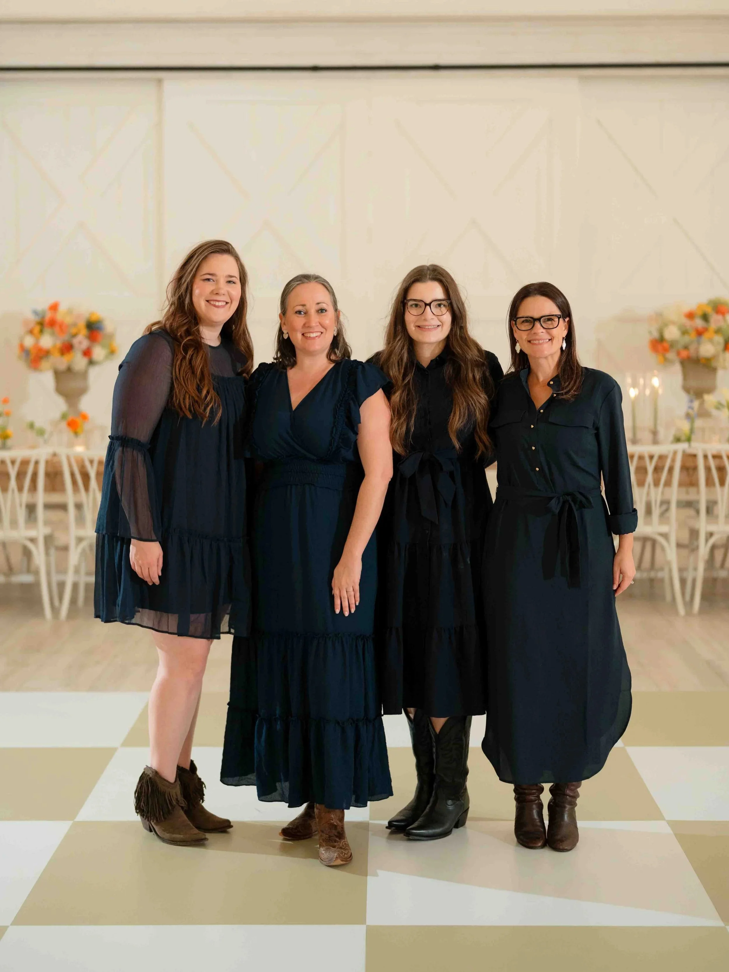 Four women in black and navy dresses standing together in a decorated event space with floral arrangements and white chairs in the background.