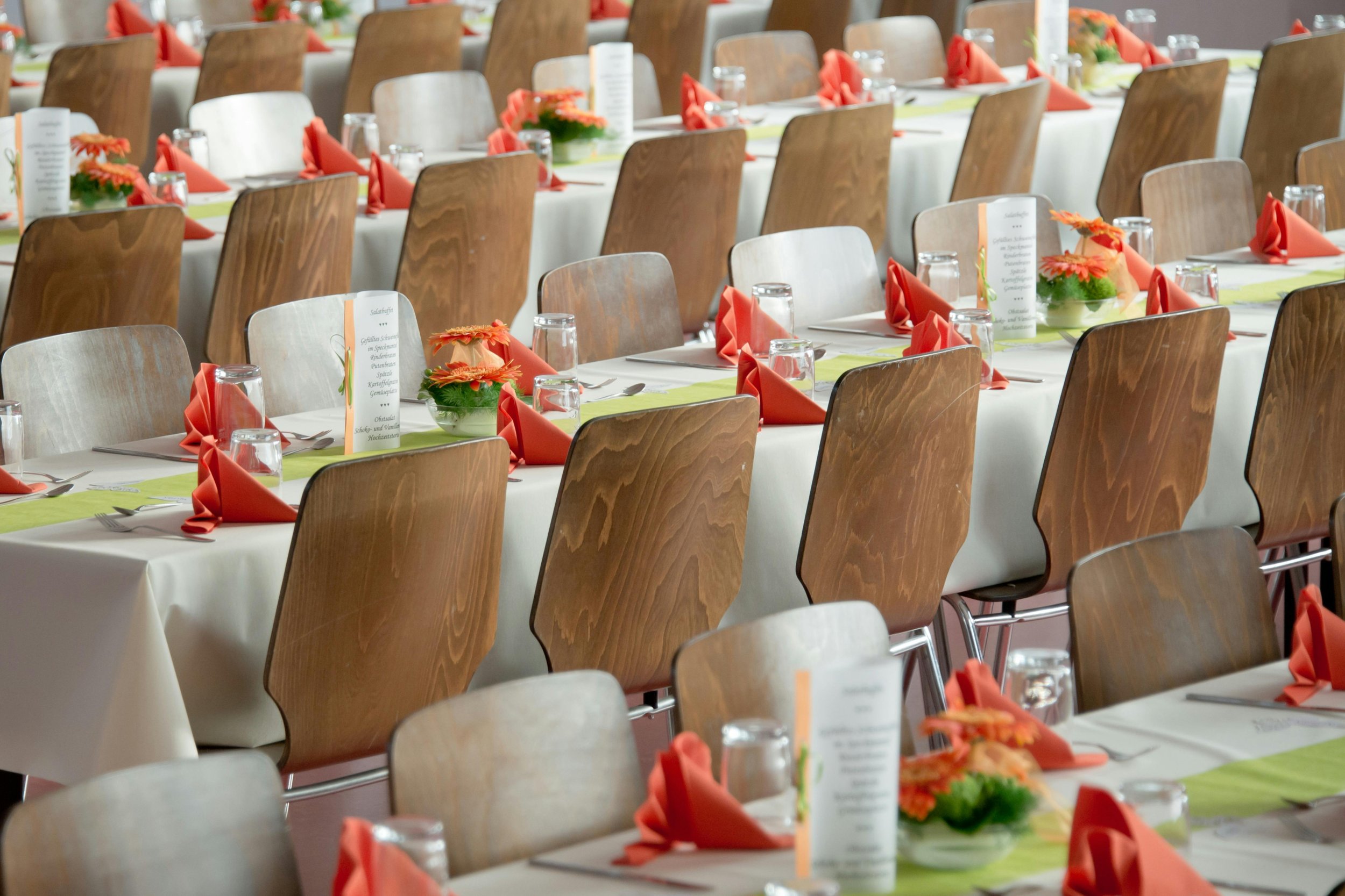 Rows of arranged dining tables with wooden chairs, set for a formal event with orange napkins folded on white tablecloths, small floral centerpieces, and printed menus.