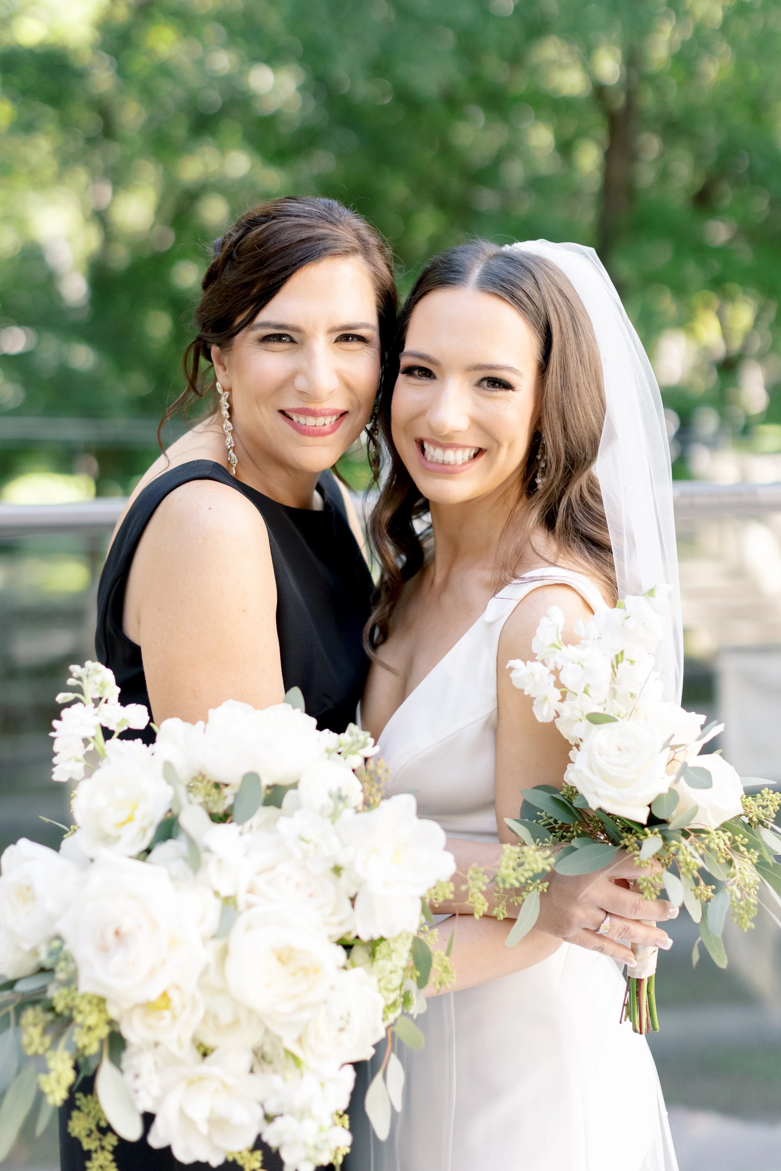 Two women, one in a white wedding dress and veil, and the other in a black dress, standing outdoors with greenery in the background, smiling and holding bouquets of white flowers.