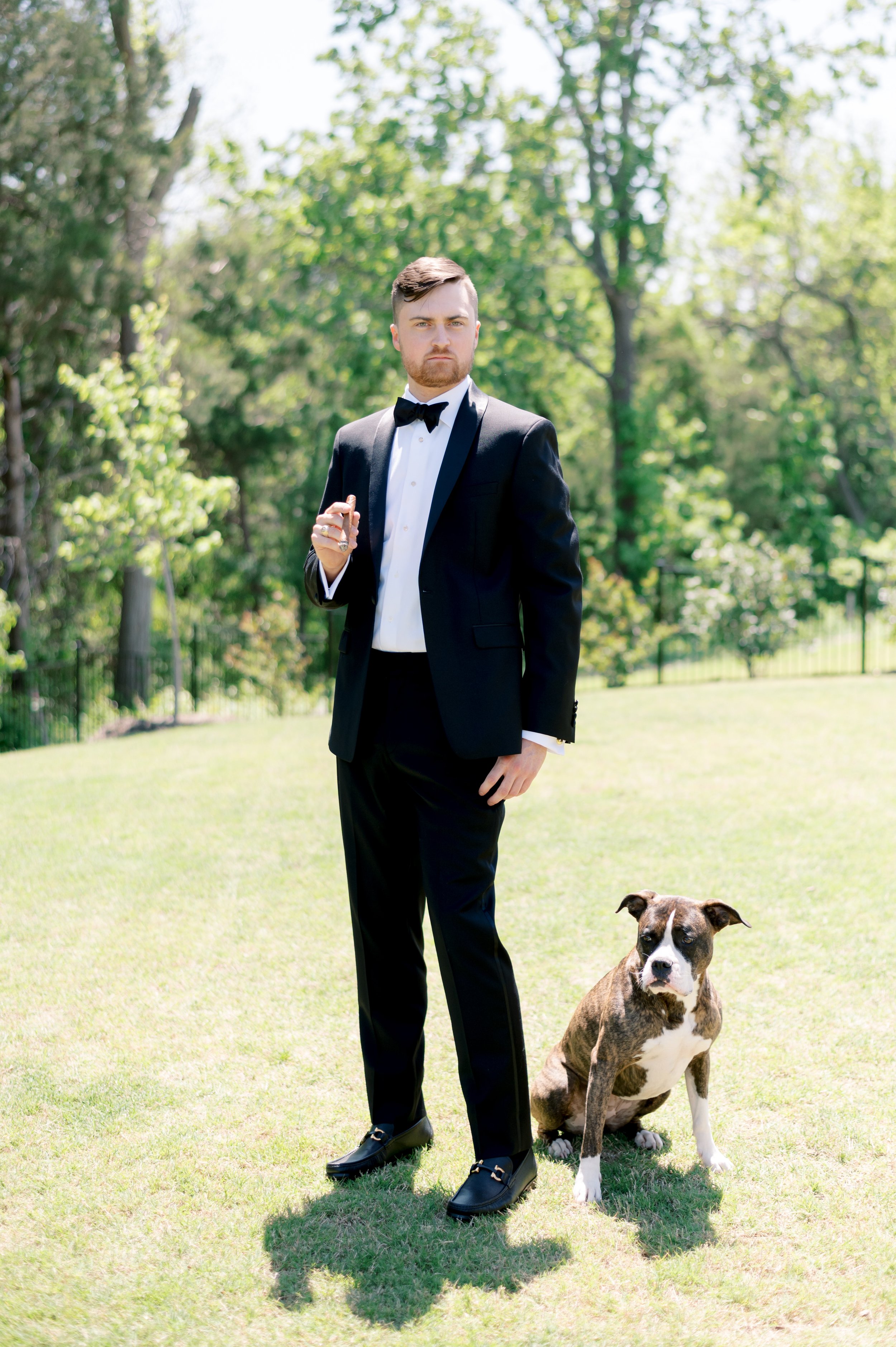 A man in a black tuxedo with a bow tie standing outdoors on grass with a brindle and white dog sitting next to him.