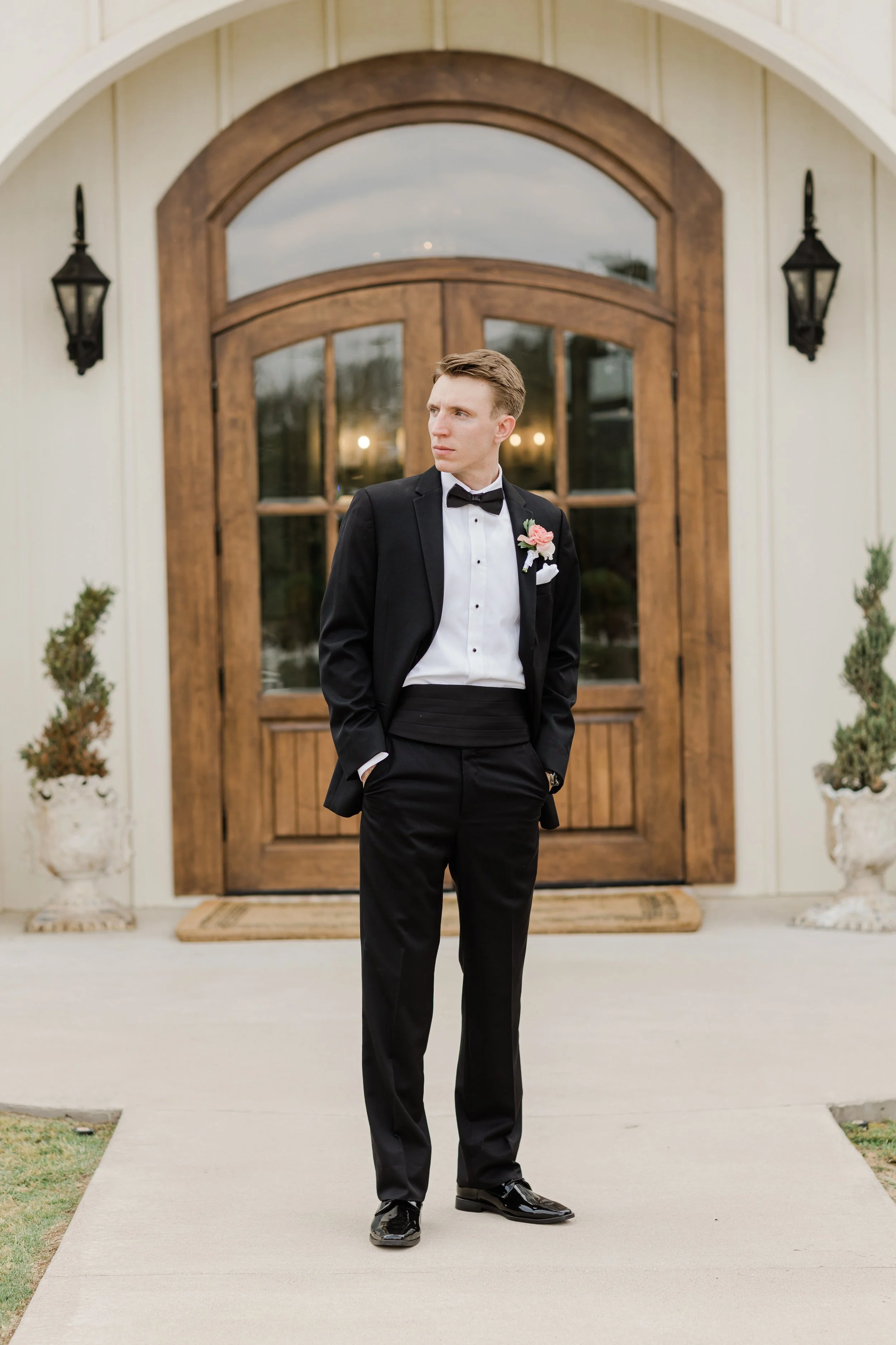 A young man in a tuxedo standing outside in front of a wooden double door with glass panels and black lantern wall sconces, with potted plants on either side.