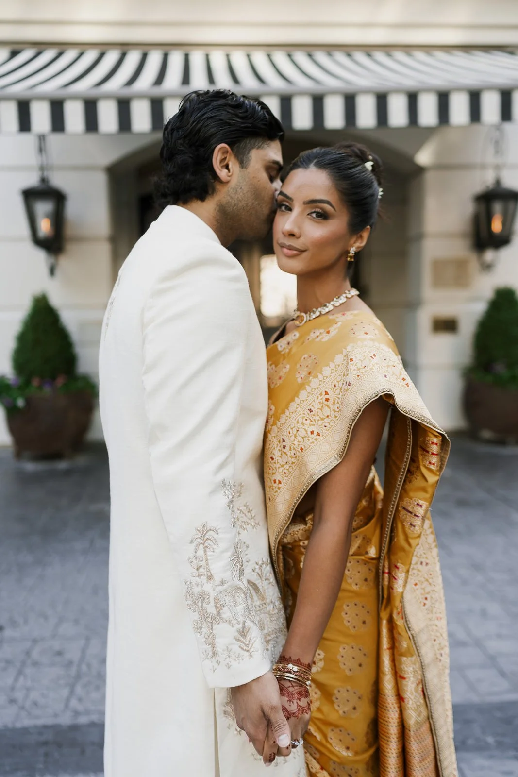 A couple dressed in traditional Indian wedding attire, standing close together outside a building with striped awning and potted plants.