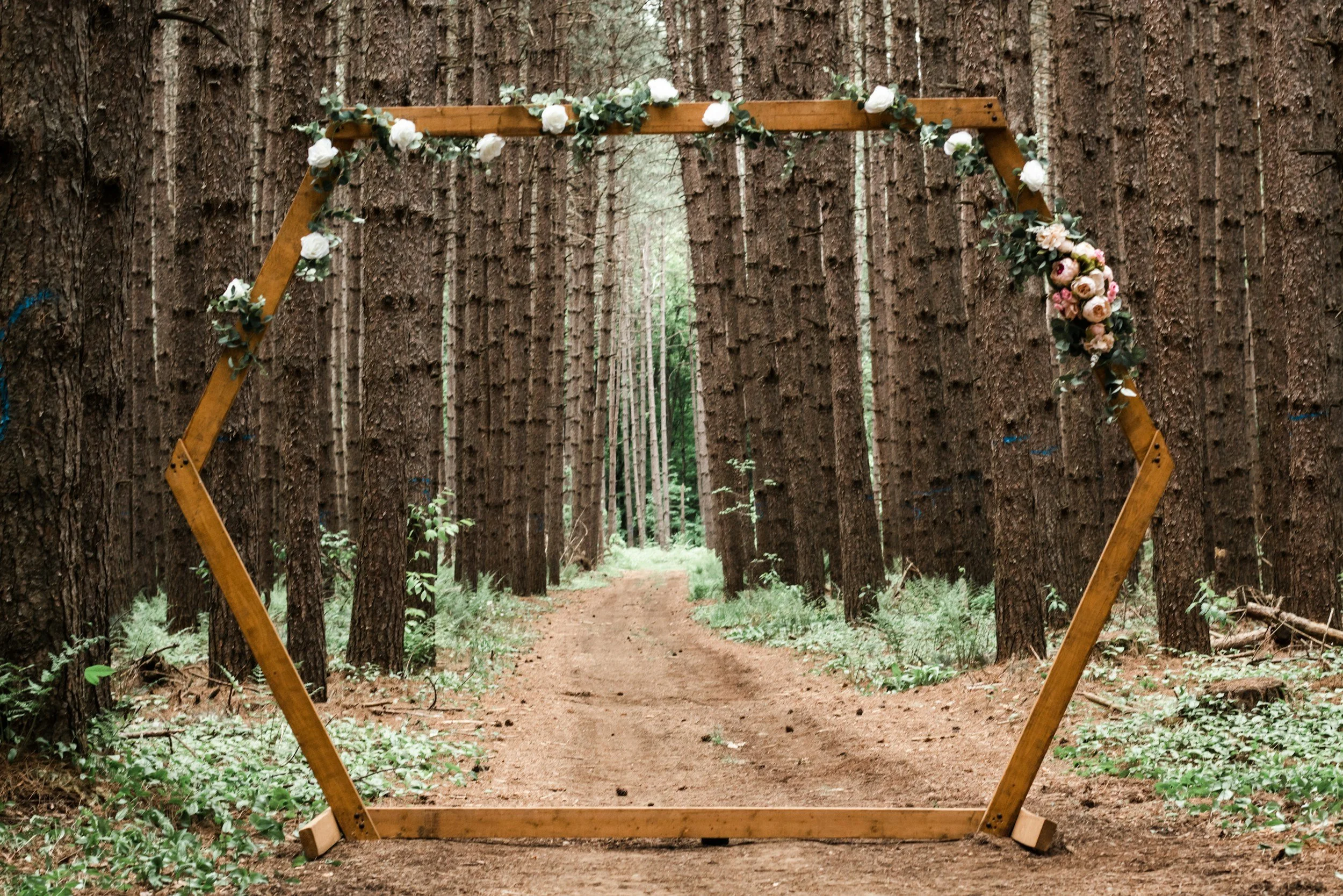 Wooden hexagonal wedding arch decorated with flowers and greenery, set in a forest with tall trees along a dirt path.