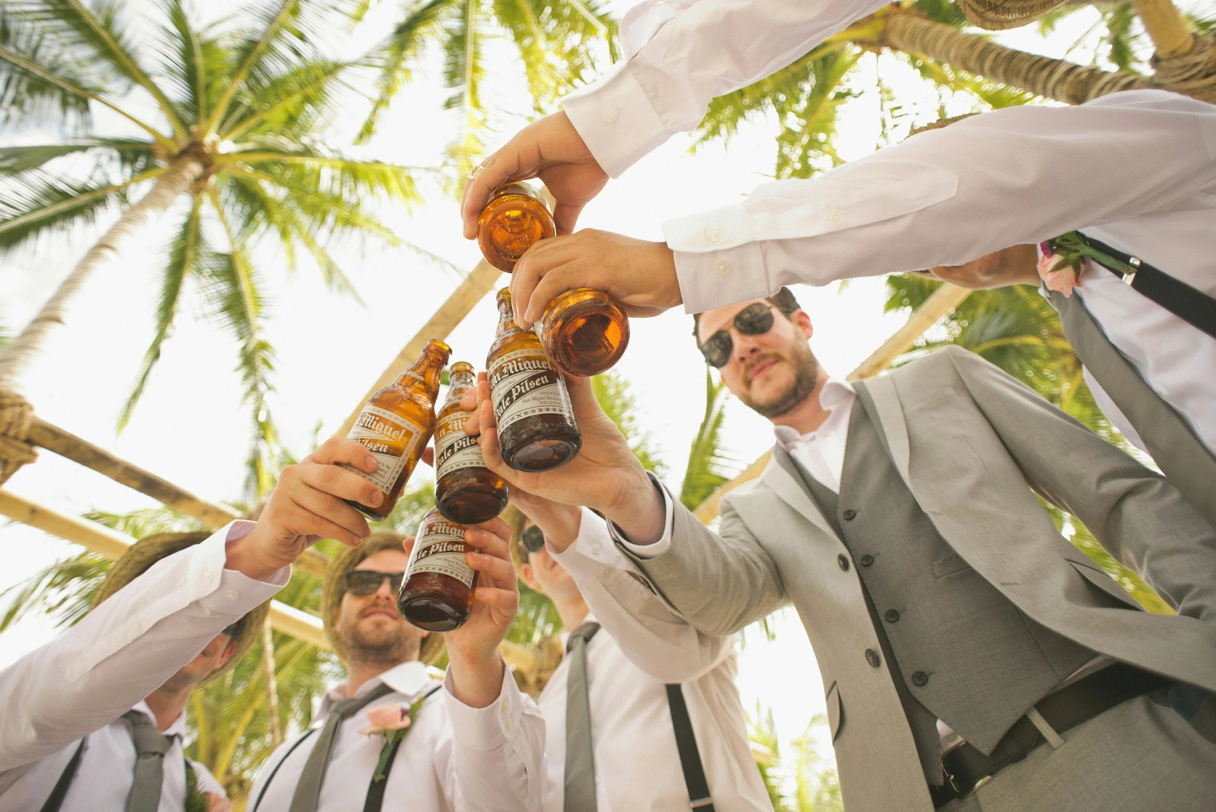 Group of men wearing suits and sunglasses raising beer bottles in a toast outdoors with palm trees.