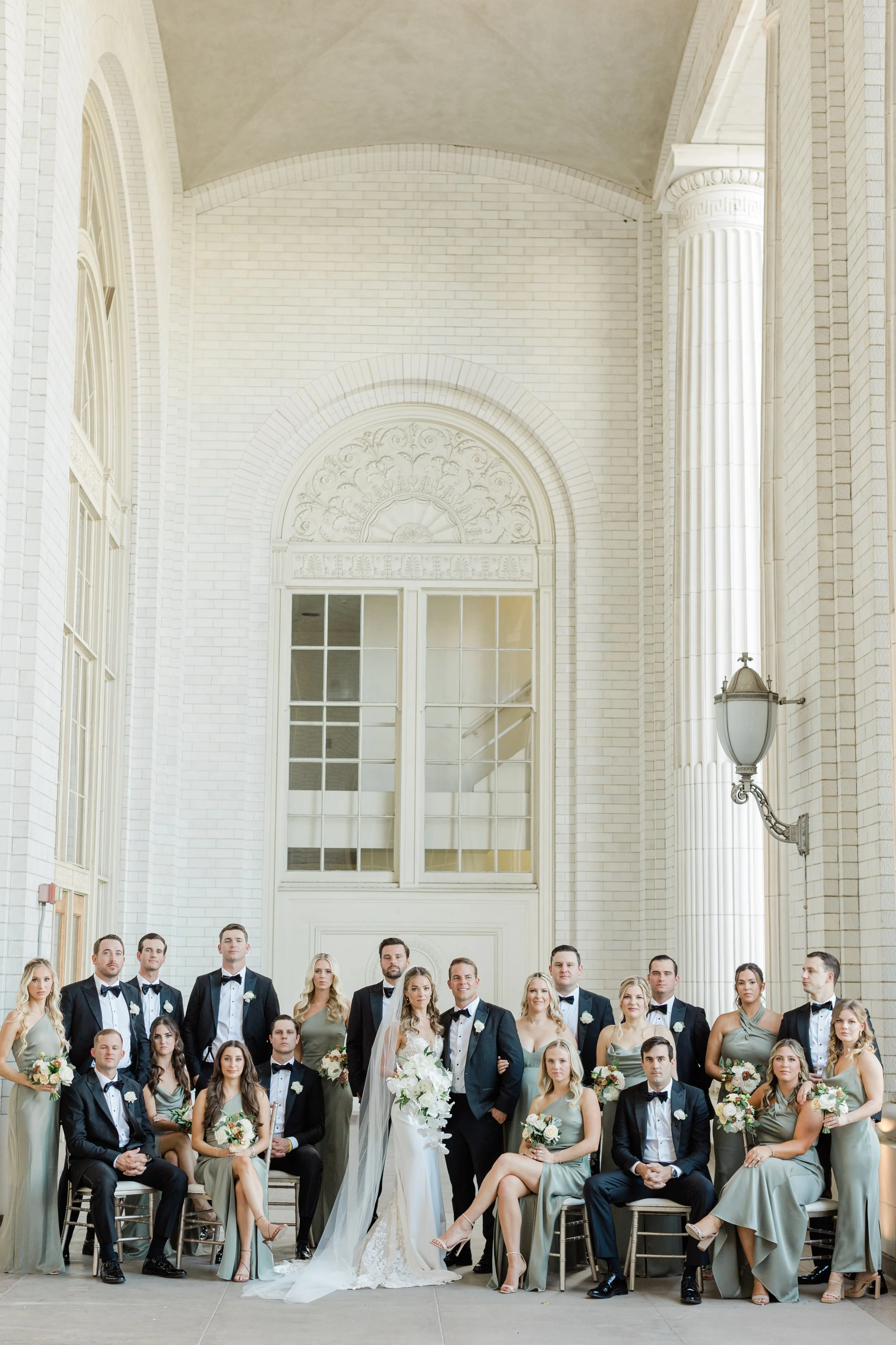 A large wedding party in formal attire, including brides and grooms, gathered inside a bright, elegant venue with high ceilings, white brick walls, and tall arched windows.