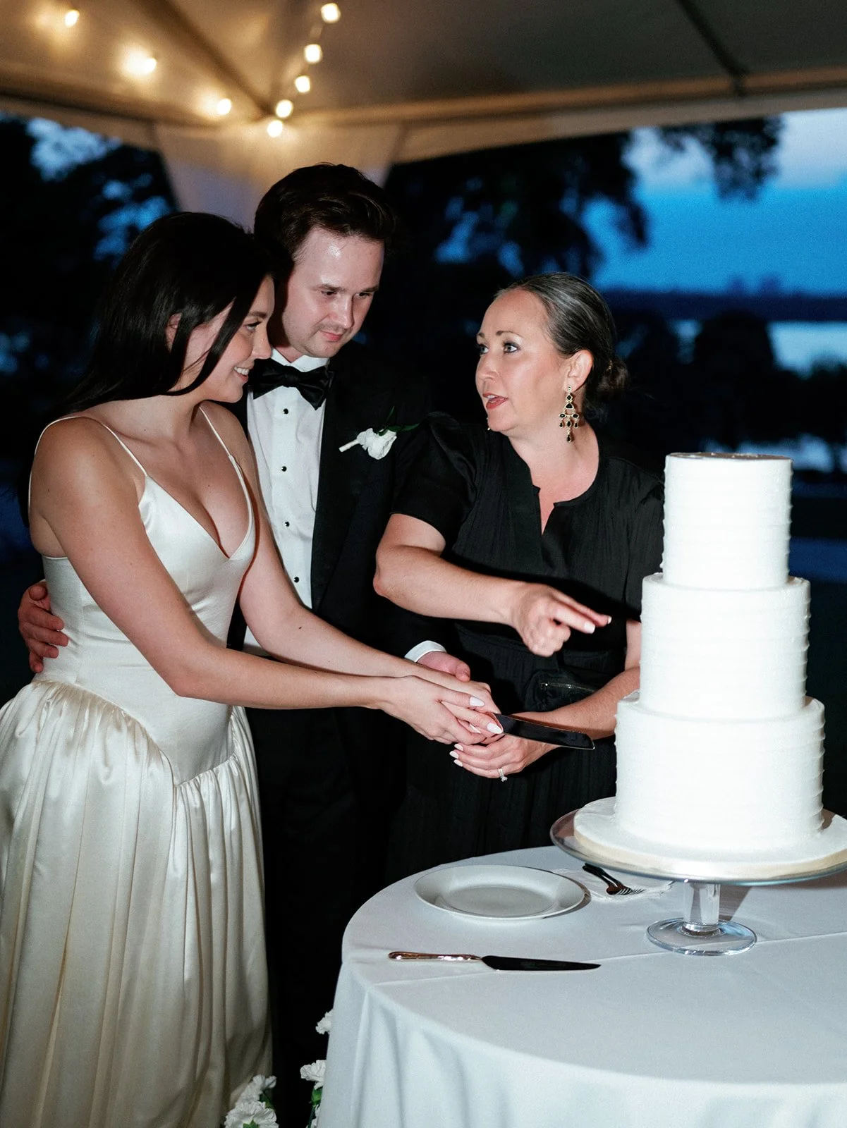 A bride and groom cutting a wedding cake with a woman standing beside them.