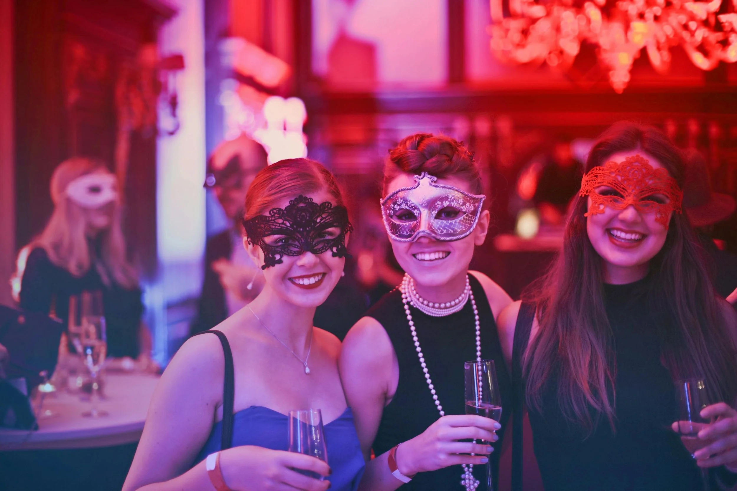 Three women smiling at a masquerade party, wearing elaborate masks and holding glasses of champagne.