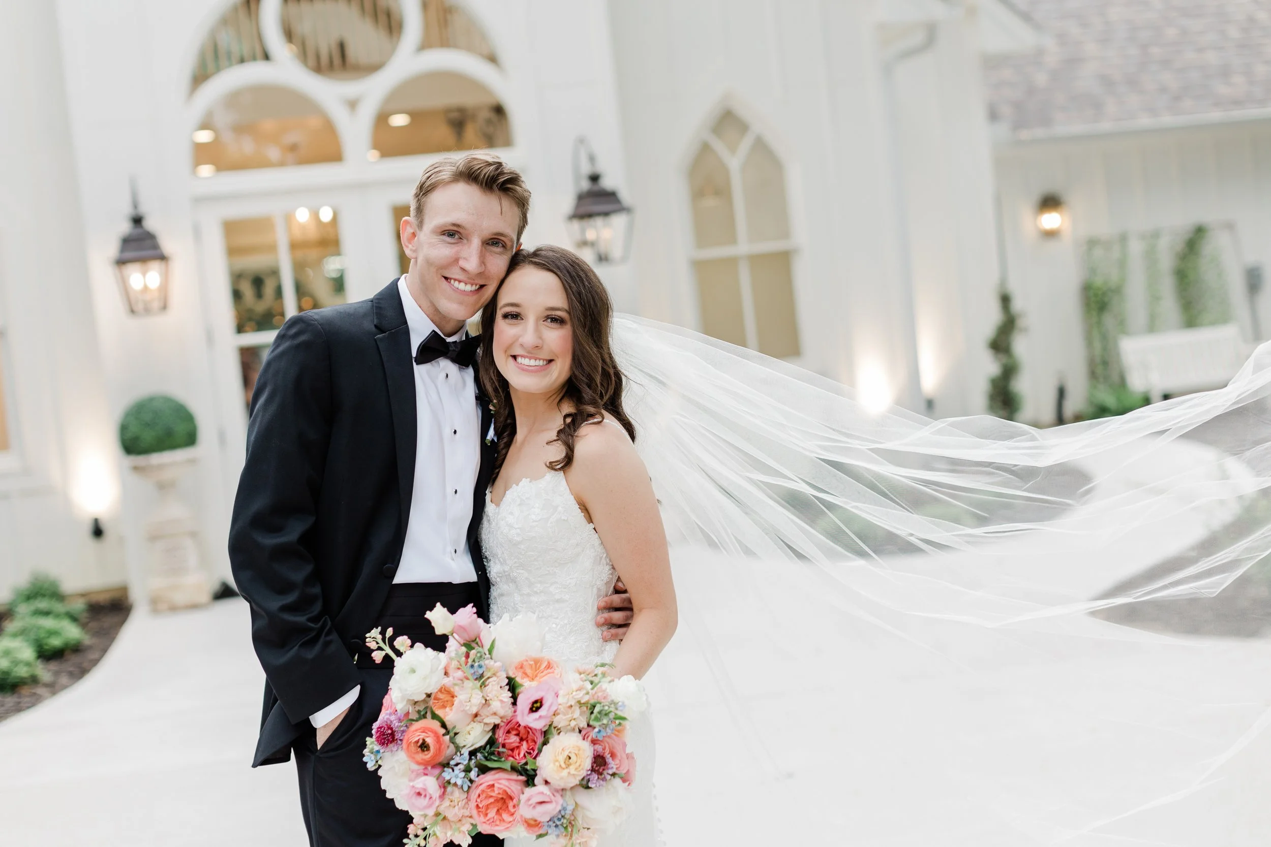 A happy bride and groom in wedding attire posing outside a building with arched windows and lanterns, holding a bouquet of pink, peach, and white flowers.