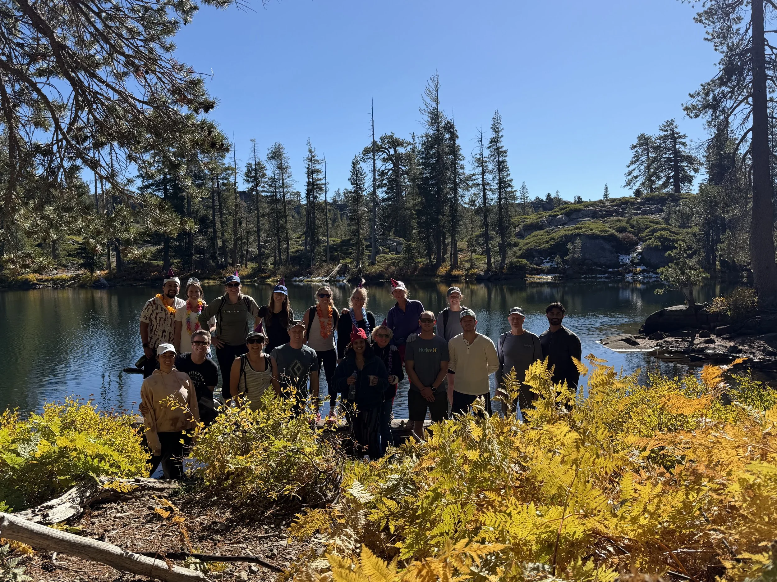 Group of about 15 people posing by a lake with trees and mountains in the background, on a sunny day.