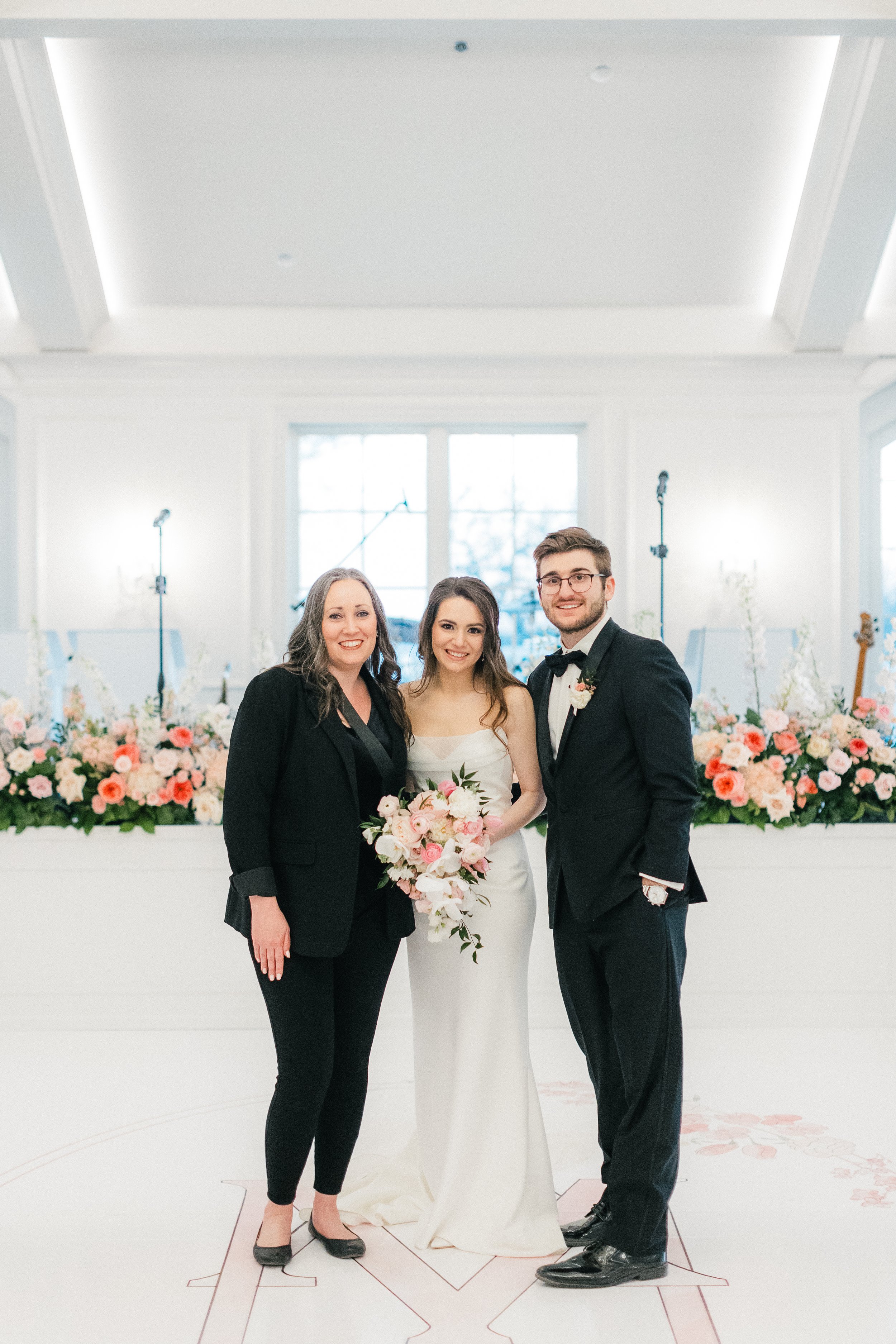 A bride and groom standing with a woman at a wedding reception, with floral arrangements and instruments in the background.