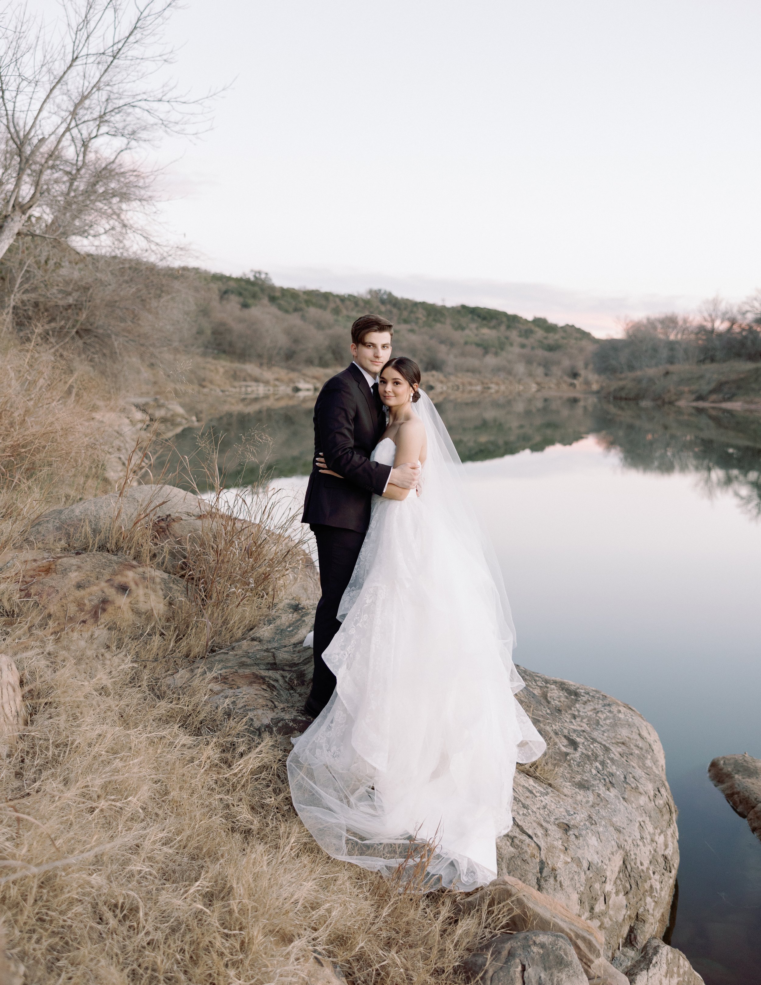A bride and groom standing on rocks by a lake during sunset, with the bride in a white wedding gown and veil and the groom in a black suit, embracing each other.