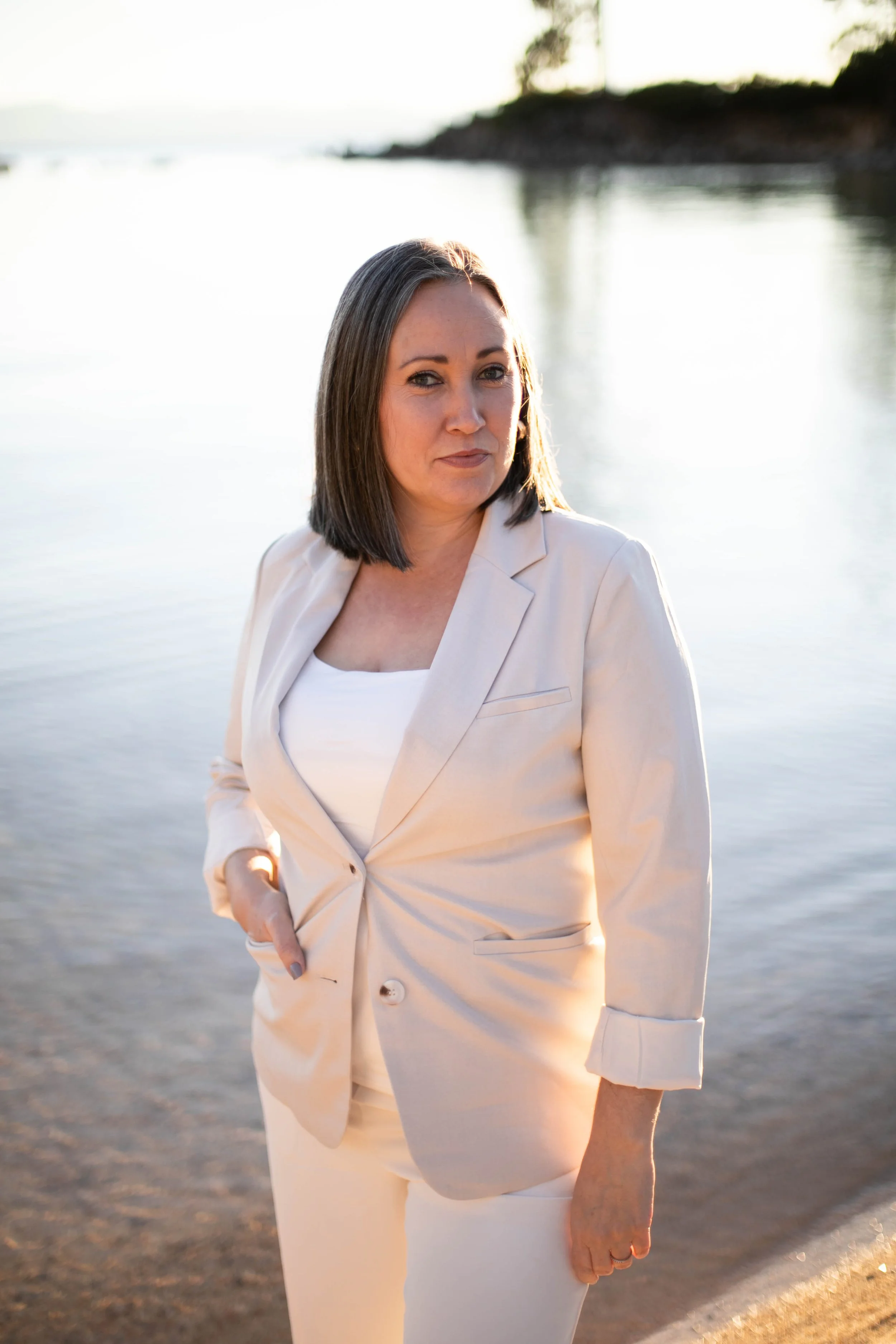 A woman with shoulder-length dark hair standing by a body of water at sunset, wearing a light-colored blazer and matching pants.