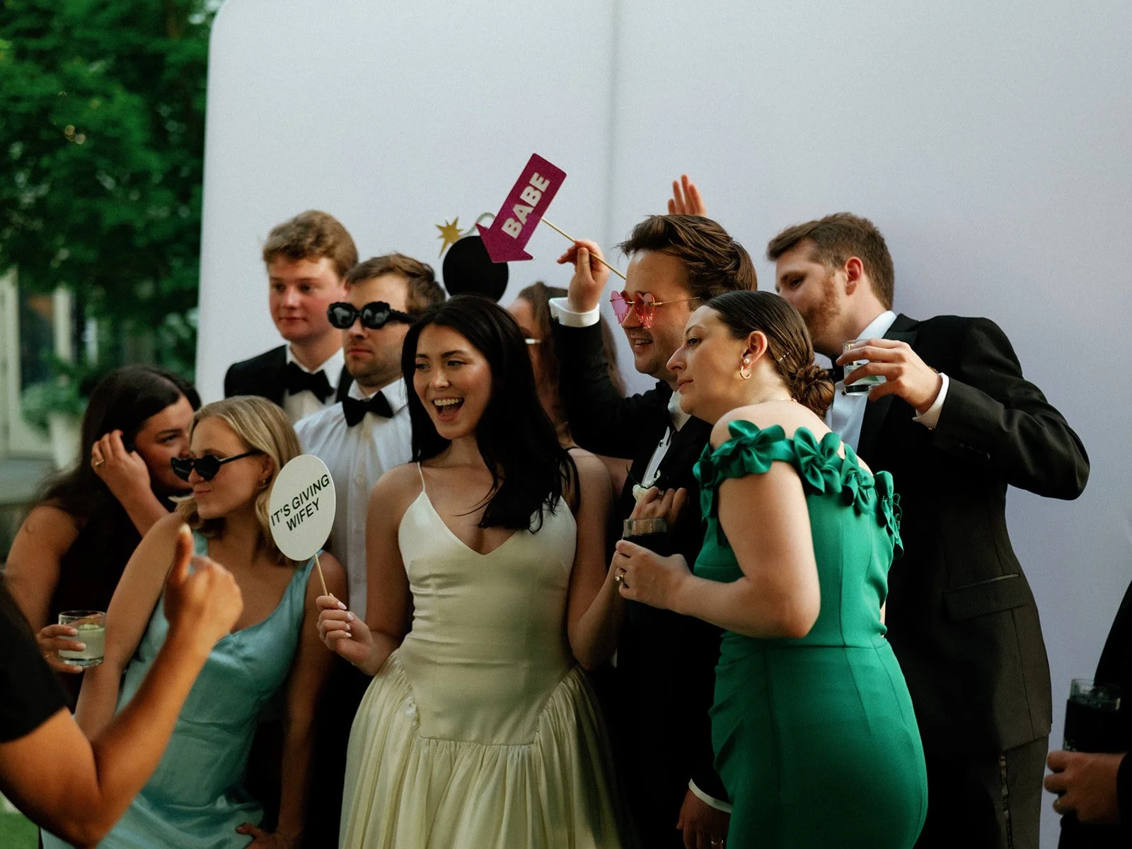 Group of people at a celebration, some holding drinks and wearing formal attire, with custom photo props such as sunglasses and signs.