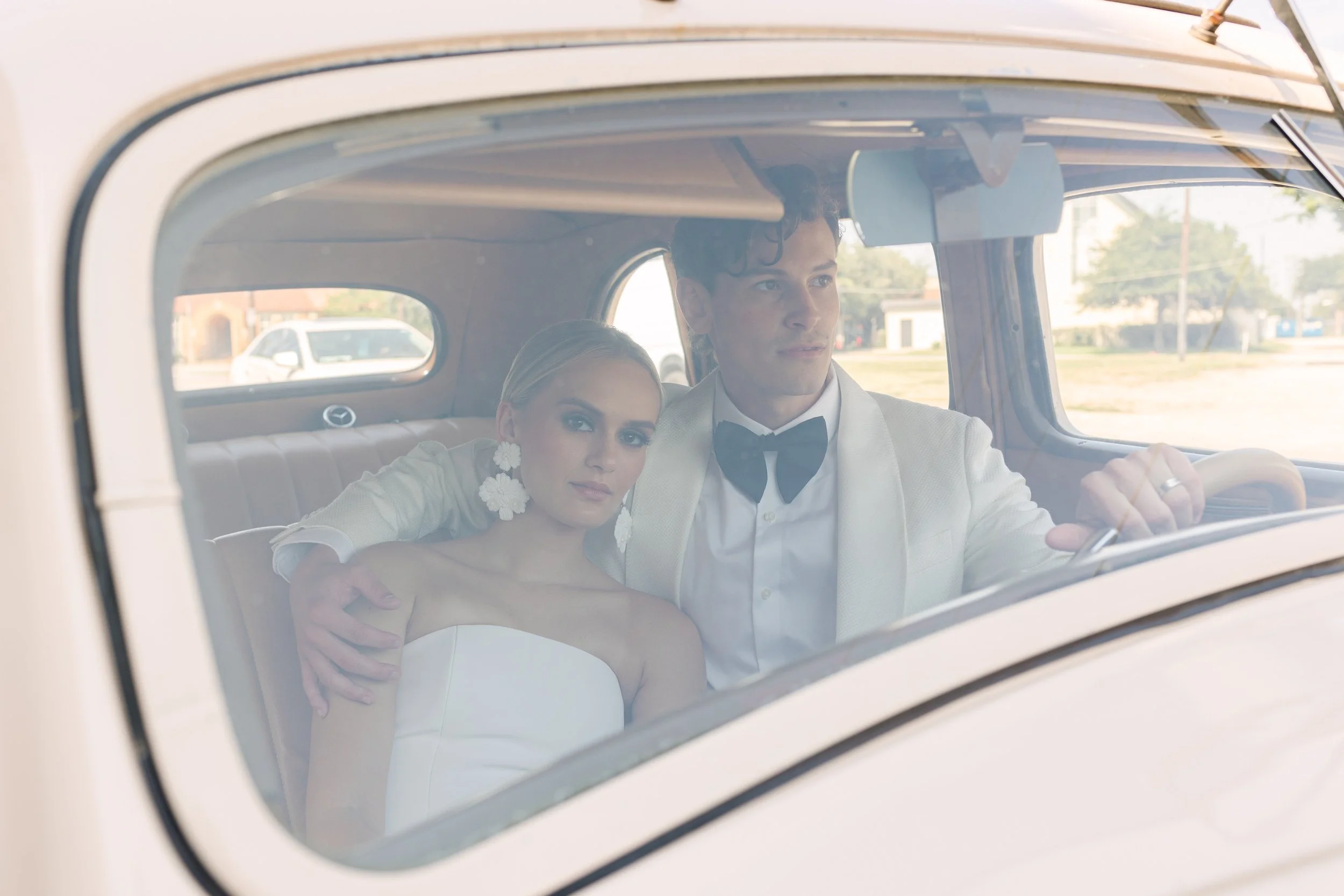 A bride and groom sitting in the backseat of a vintage car, with the bride leaning against the groom and both looking forward.