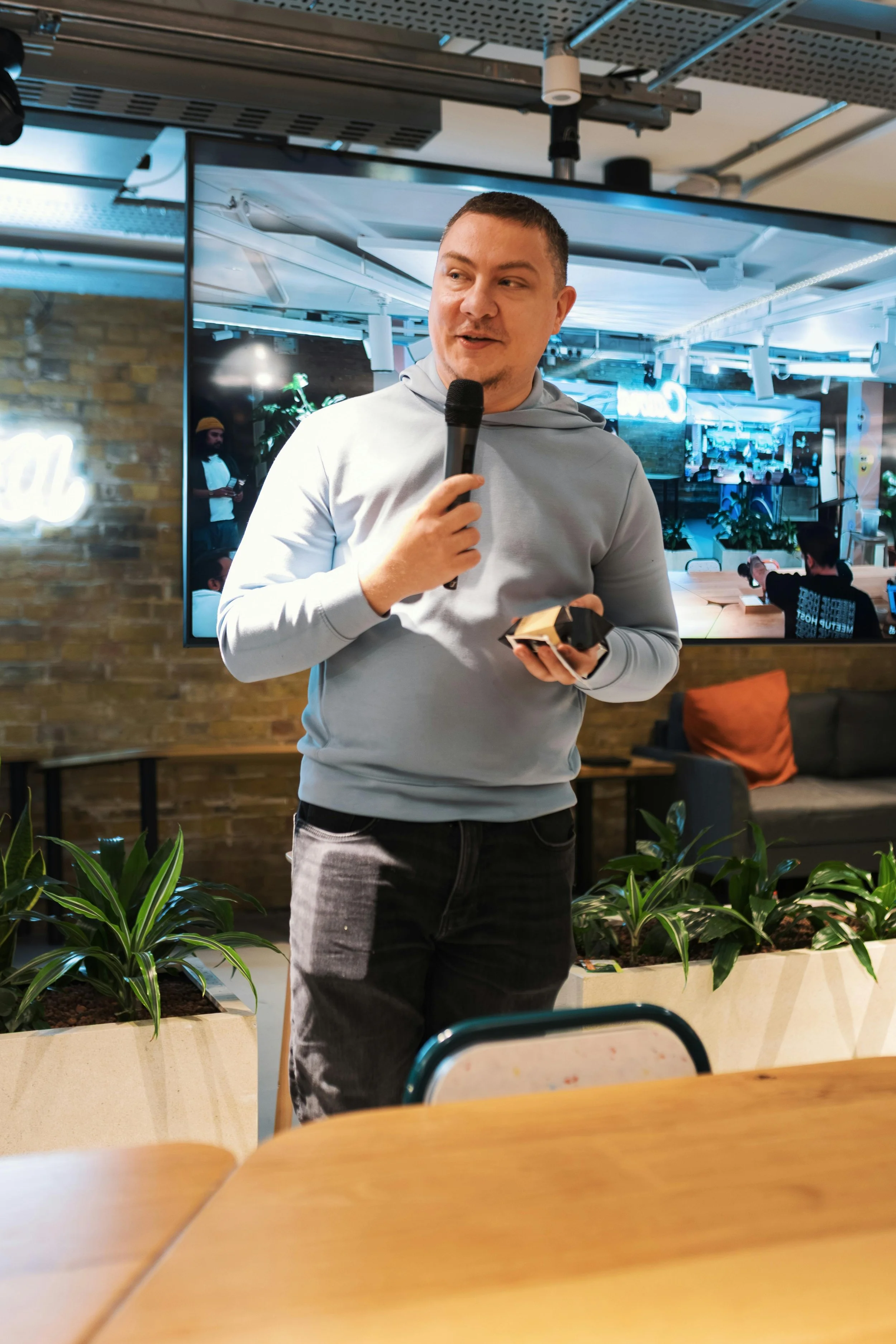 A man in a light gray hoodie and dark pants holding a microphone and a small object, possibly a card or a phone, while speaking at an indoor event or gathering. Behind him is a large television screen displaying a blue-toned room with people and plants, and the setting features a brick wall, indoor plants, and casual furniture.