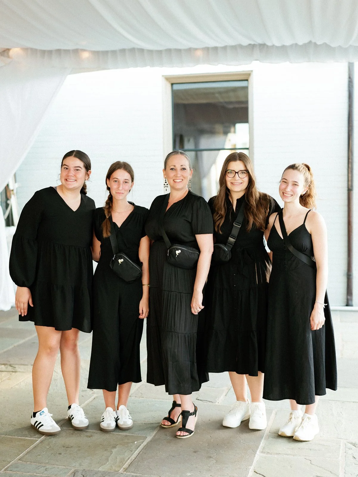 Five women in black dresses and sneakers or sandals standing together indoors, smiling at the camera.
