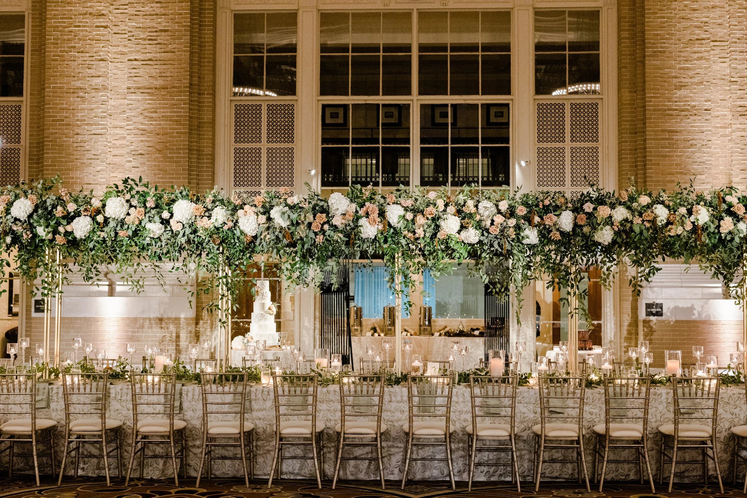 Elegant banquet table decorated with candles, greenery, and pink and white flowers, set against a brick wall with large windows at an indoor event or wedding reception.