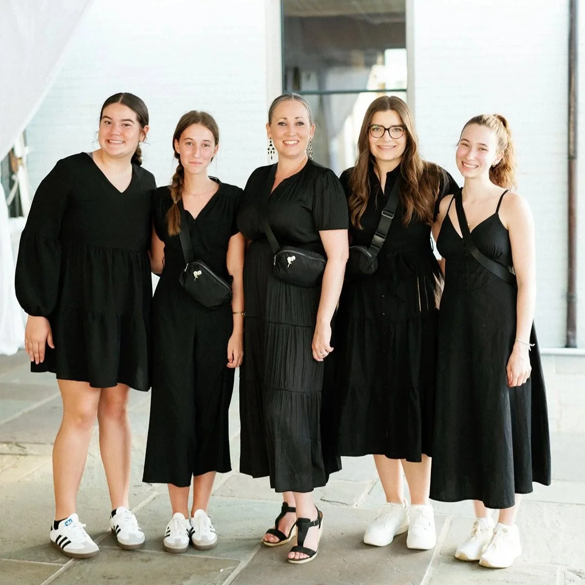 Group of five women standing together, wearing black dresses and smiling, indoors with white brick walls.