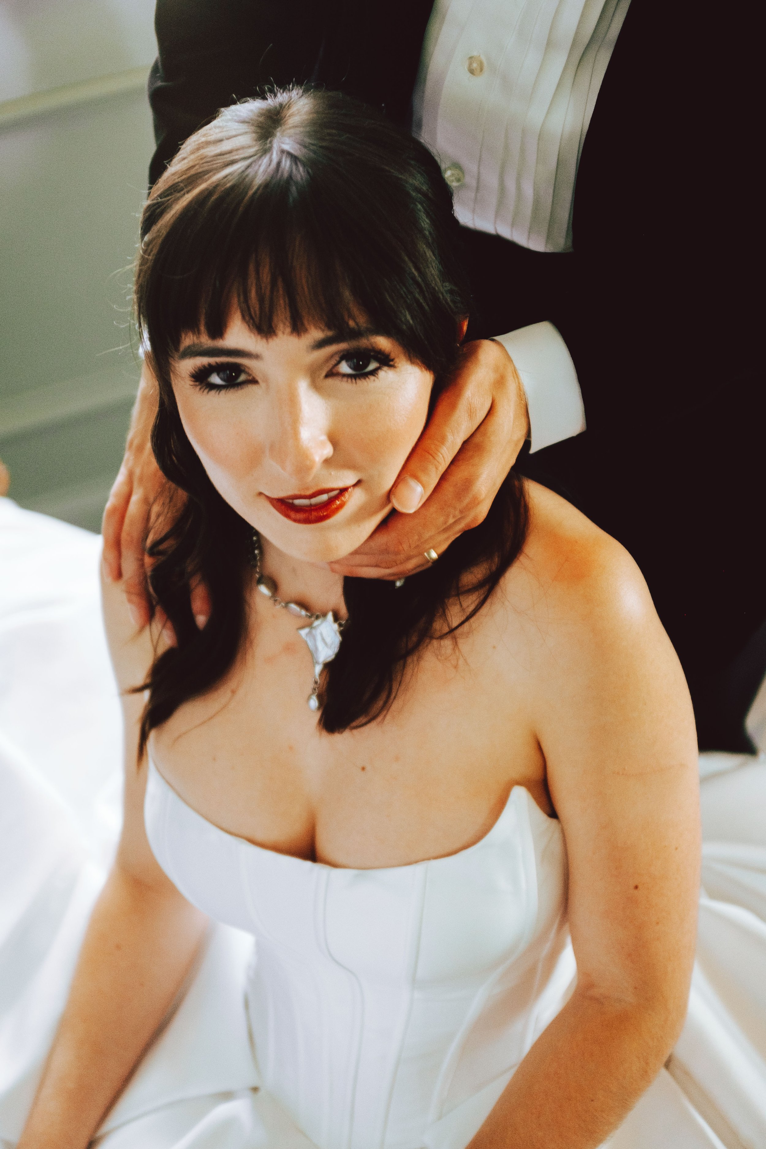 A woman in a white strapless dress with dark hair and makeup sitting on a bed, with a person in formal attire behind her.