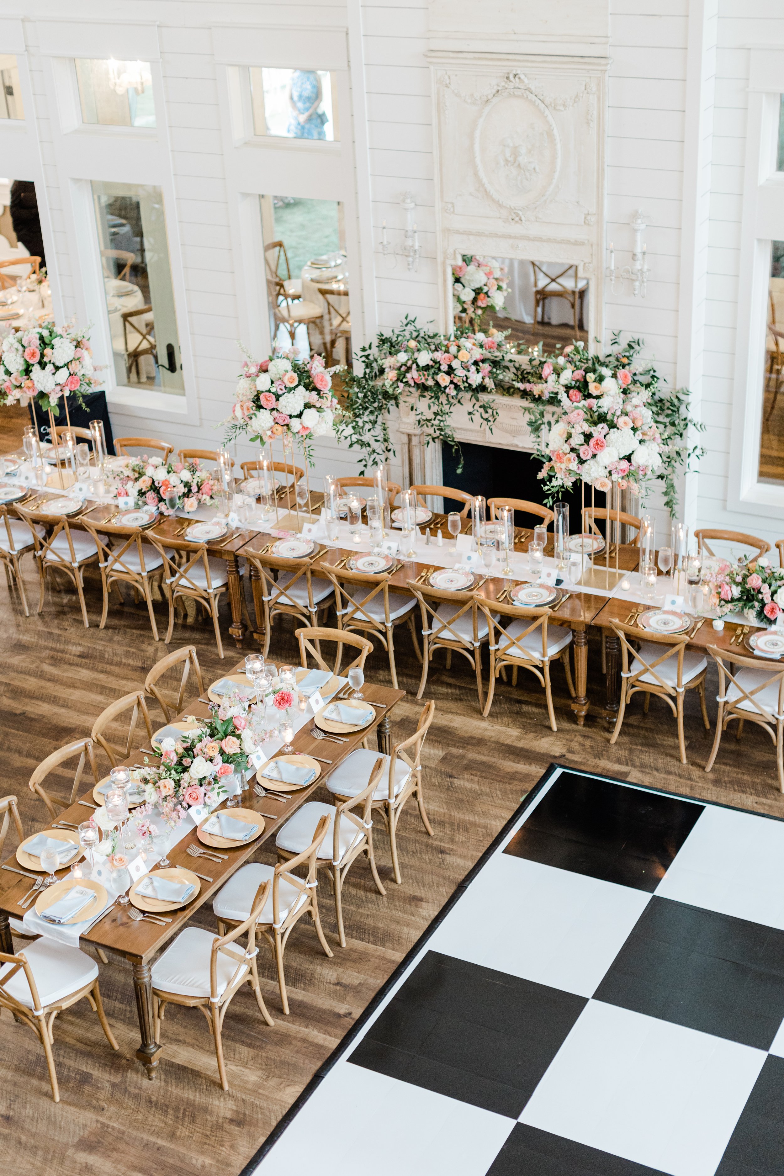 Wedding reception table decorated with pink and white flowers, candleholders, and elegant place settings, with a fireplace and large floral arrangements in a bright, white-paneled room.